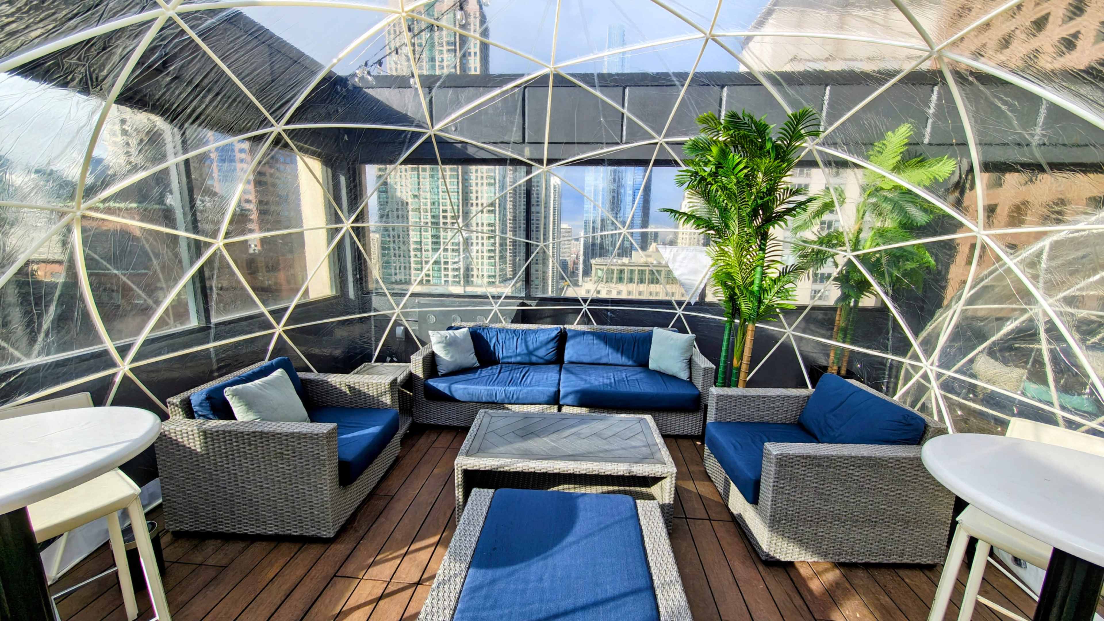 The image shows a cozy seating area inside a geodesic dome featuring wicker furniture with blue cushions, situated on a rooftop with a city skyline view.