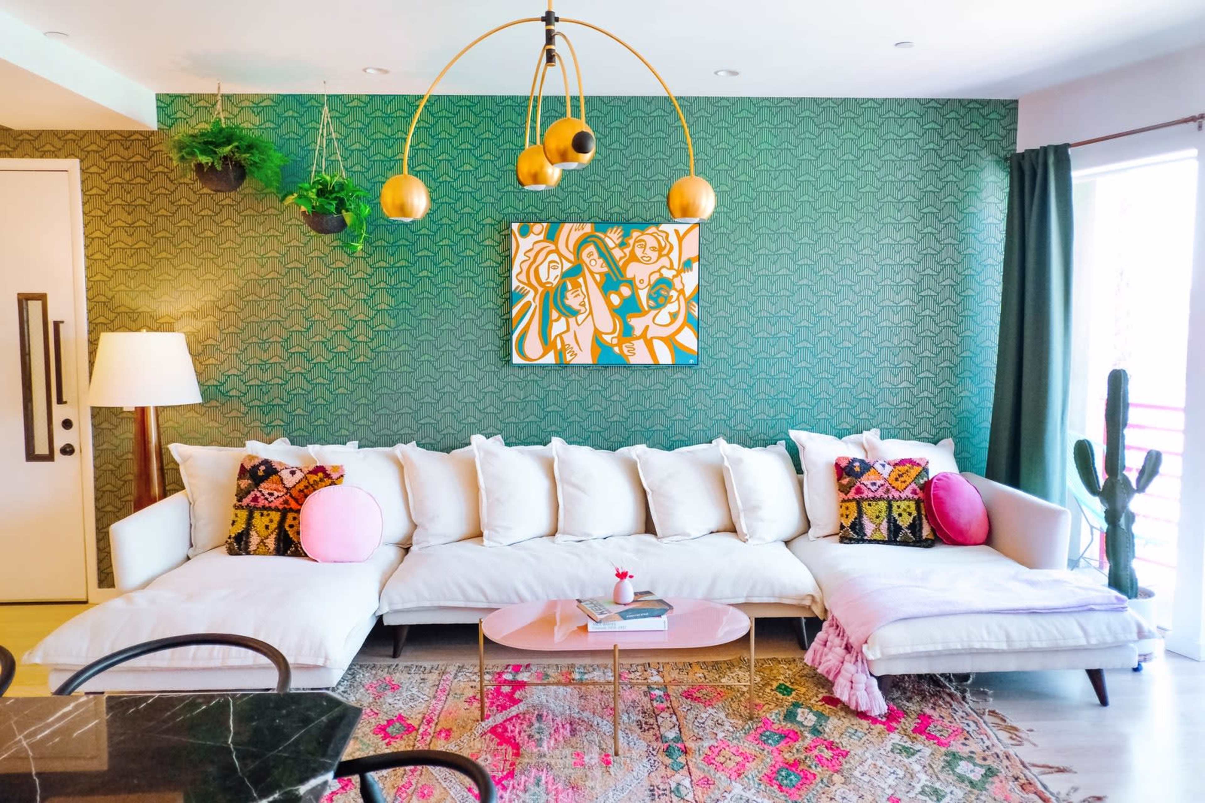 A modern living room features a large white sectional sofa adorned with colorful patterned pillows, a round pink coffee table, and vibrant artwork on the wall, set against a textured green wallpaper.