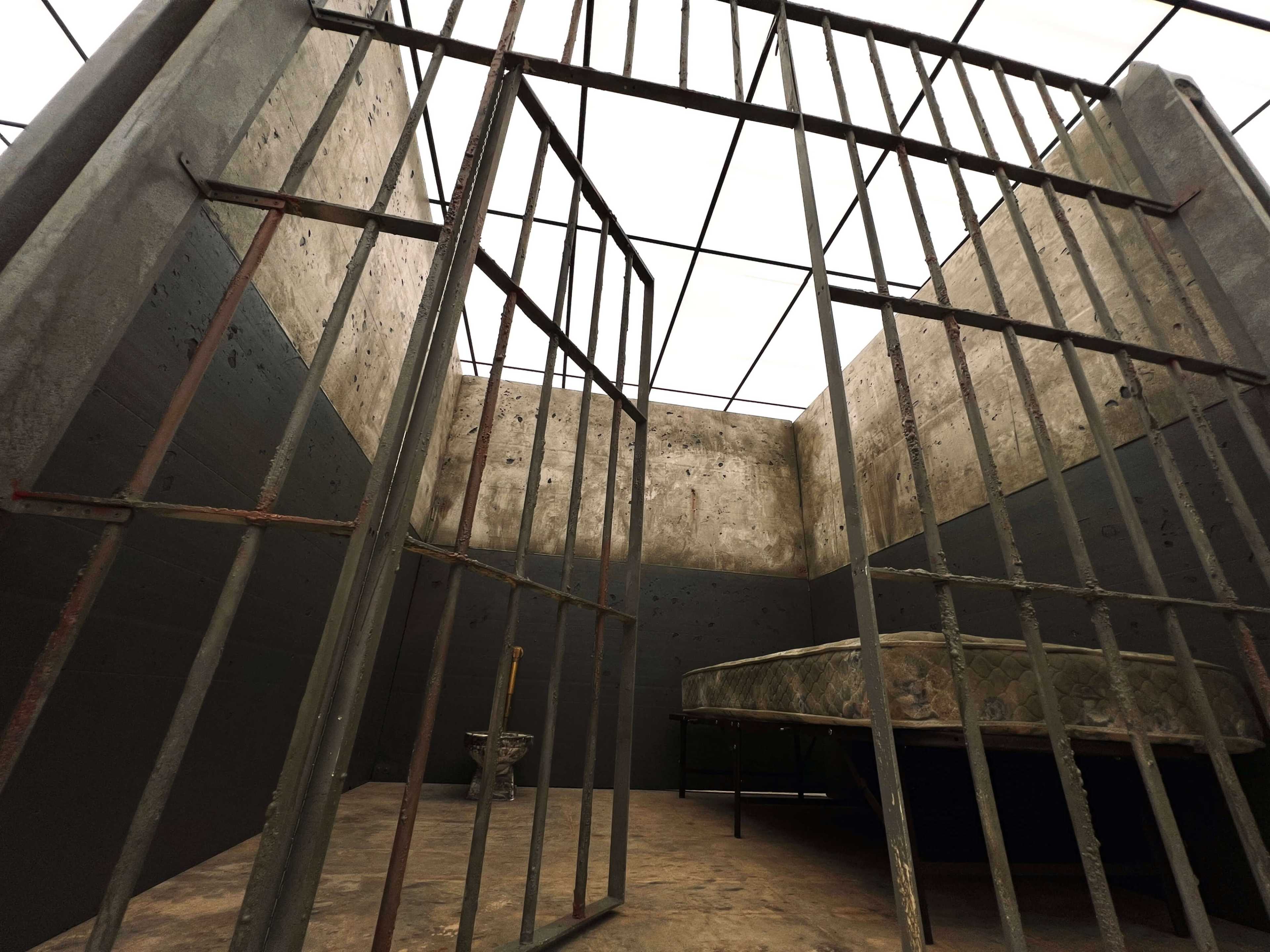 The image shows an empty prison cell with barred gates, a simple mattress on a frame, and a small pedestal sink.