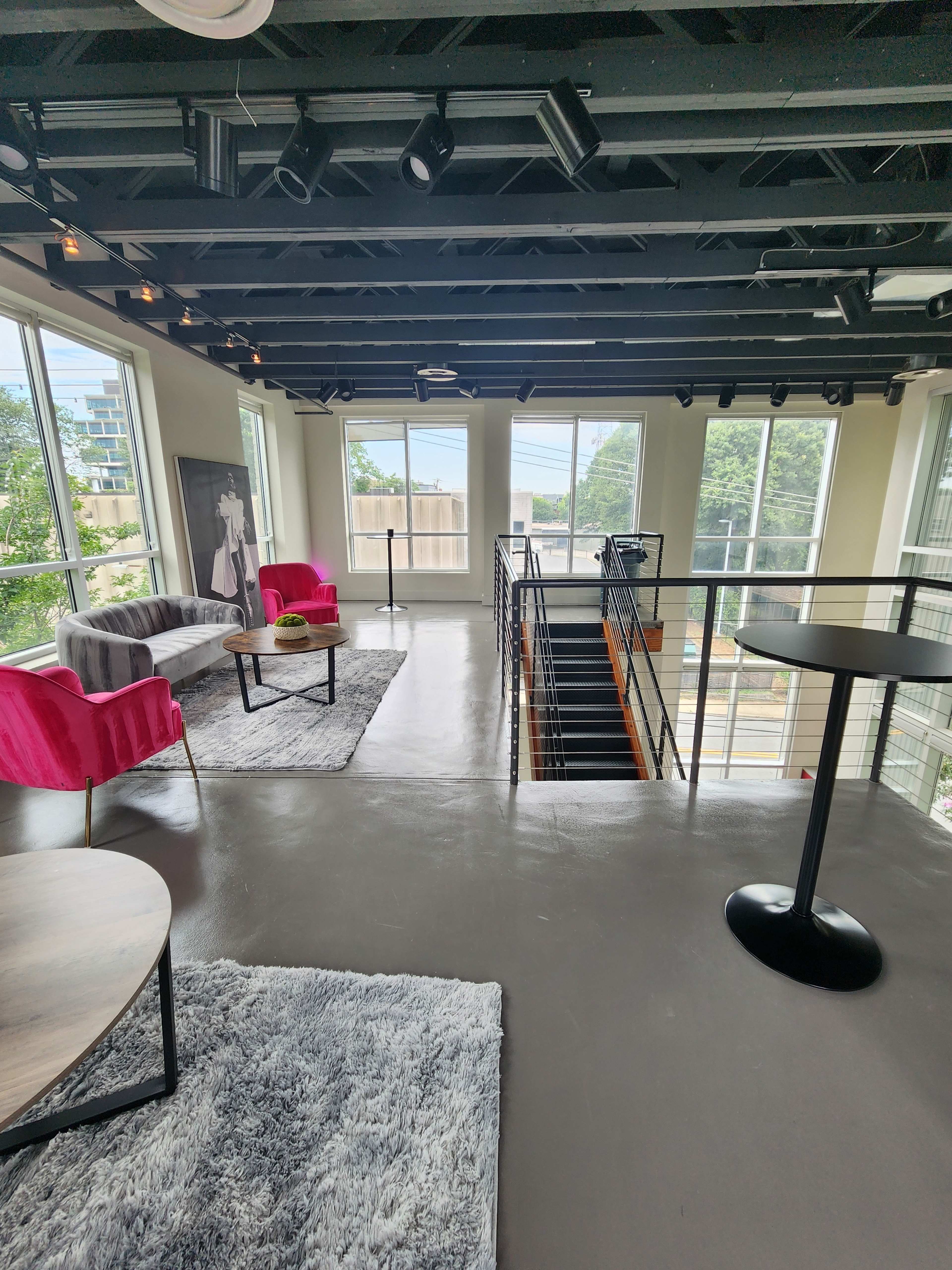 A modern interior space with pink chairs, a coffee table, and a staircase leading down, illuminated by natural light from large windows.