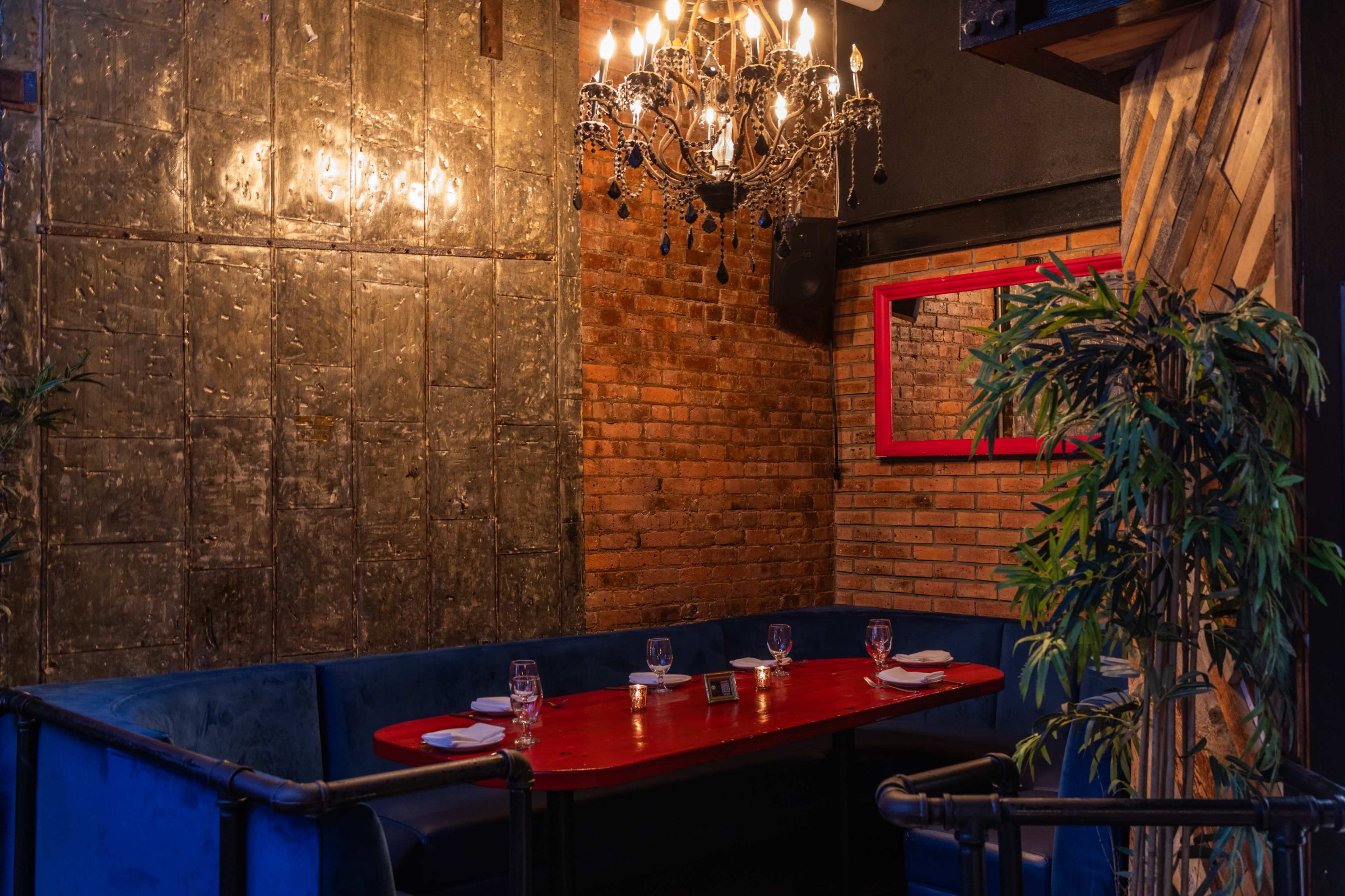 The image shows a cozy dining area with a red table set for four, surrounded by blue upholstered seating, against a backdrop of exposed brick walls and a decorative chandelier.