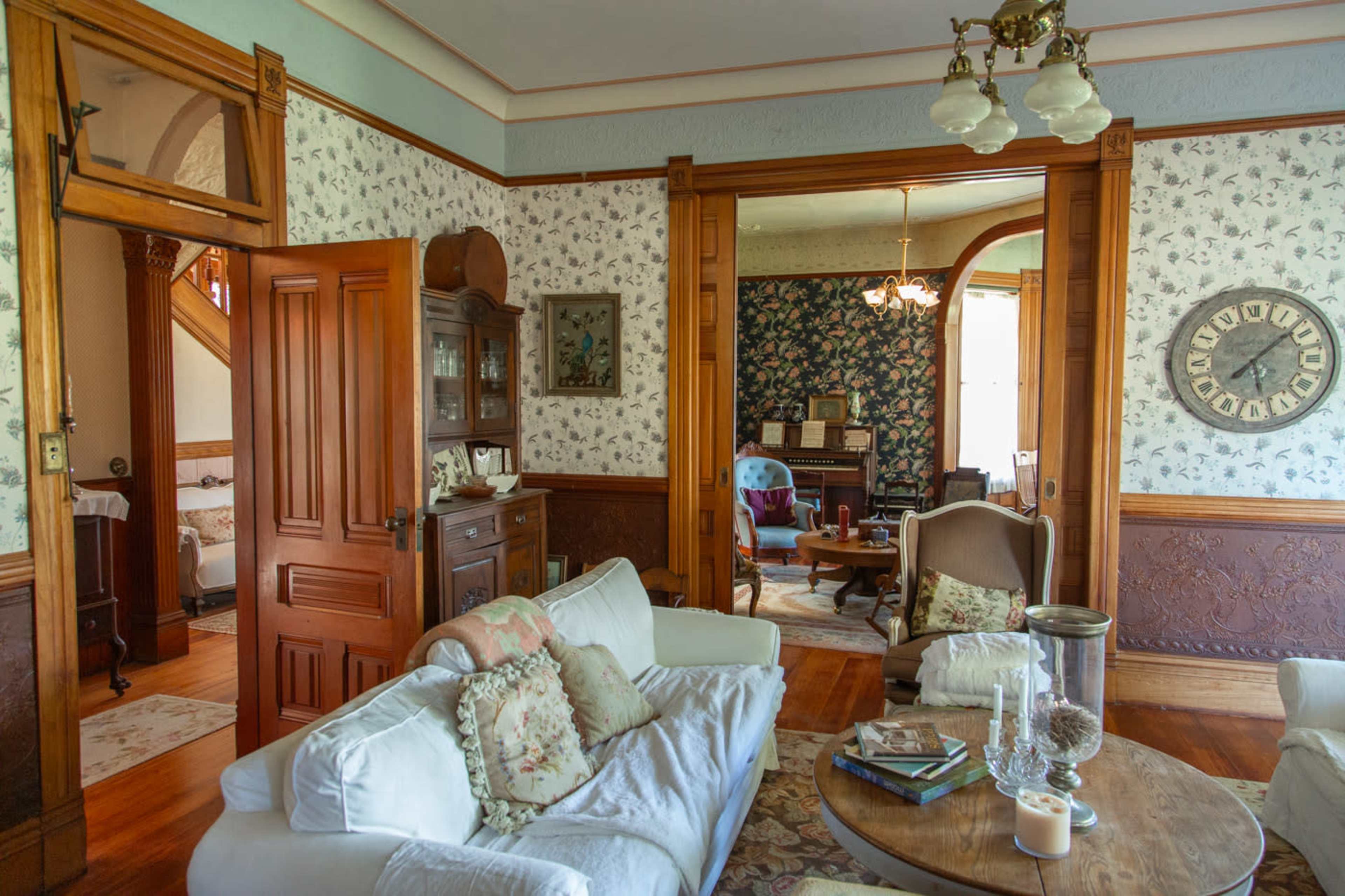 The image shows a vintage living room with floral wallpaper, a wooden dining area, and a cozy seating arrangement.