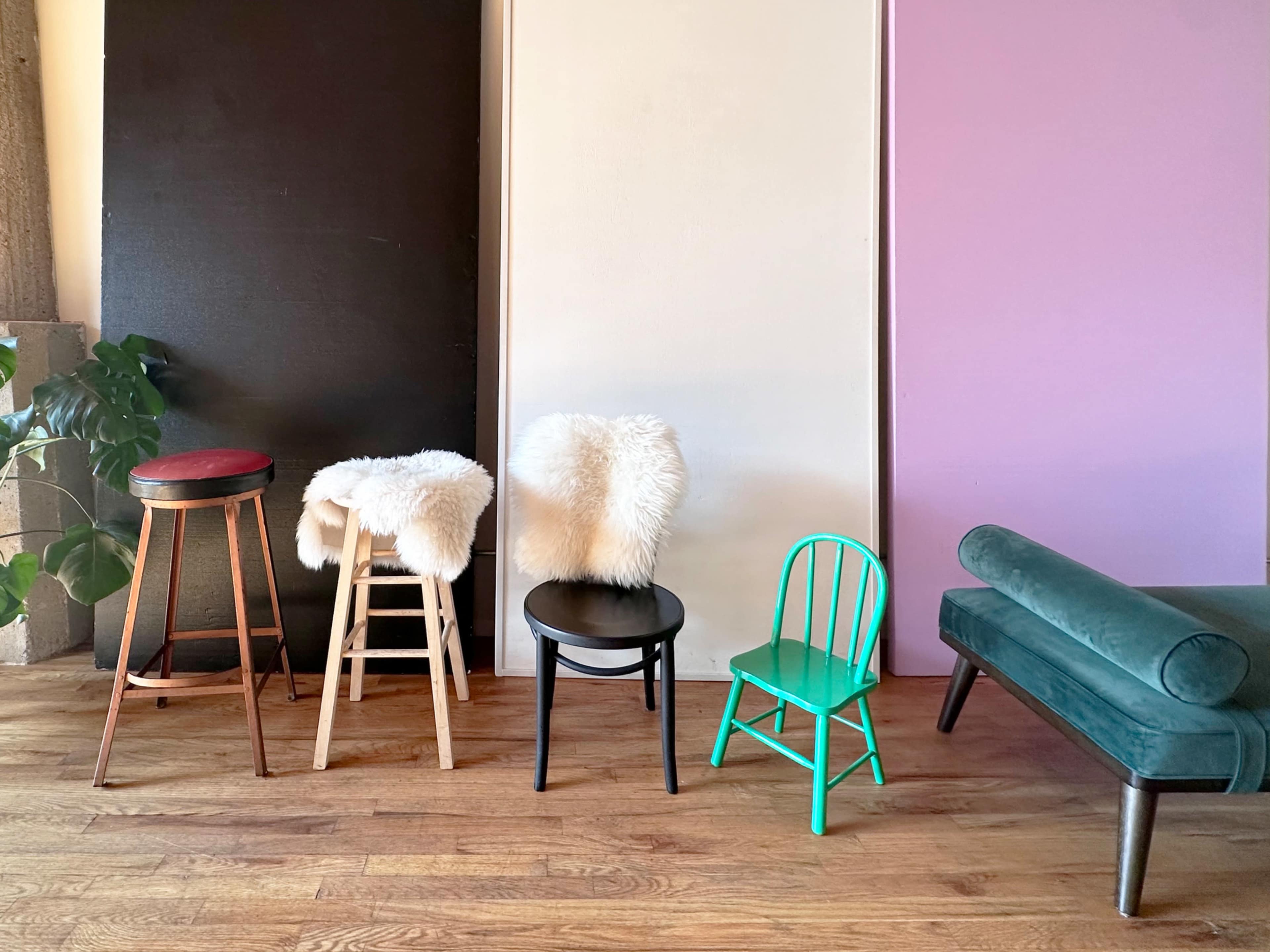 The image features a row of five chairs and stools, each with different designs, placed on a wooden floor in front of colorful backdrop panels.