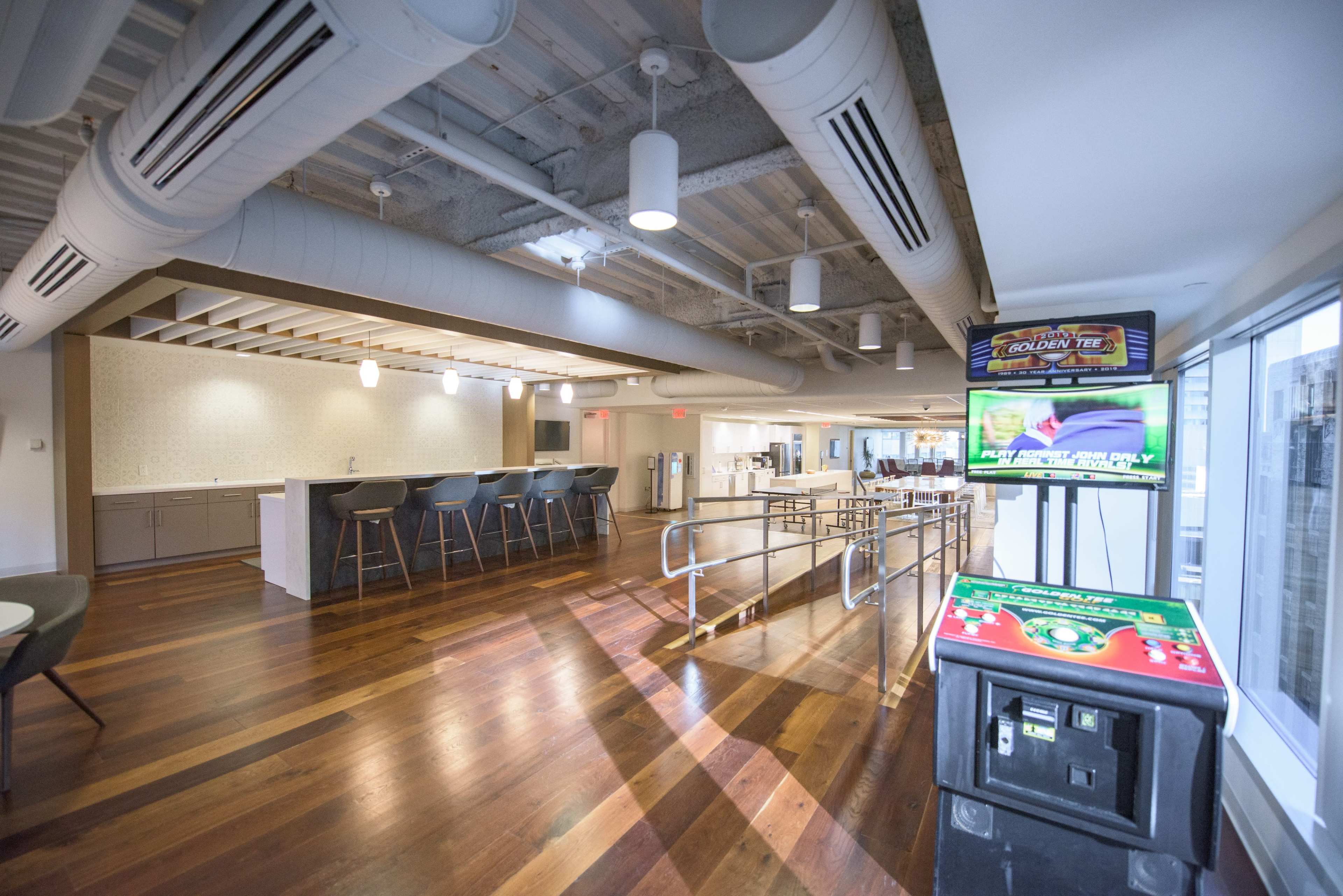A spacious modern office interior with wooden flooring, a kitchenette area, and a gaming machine in the corner.