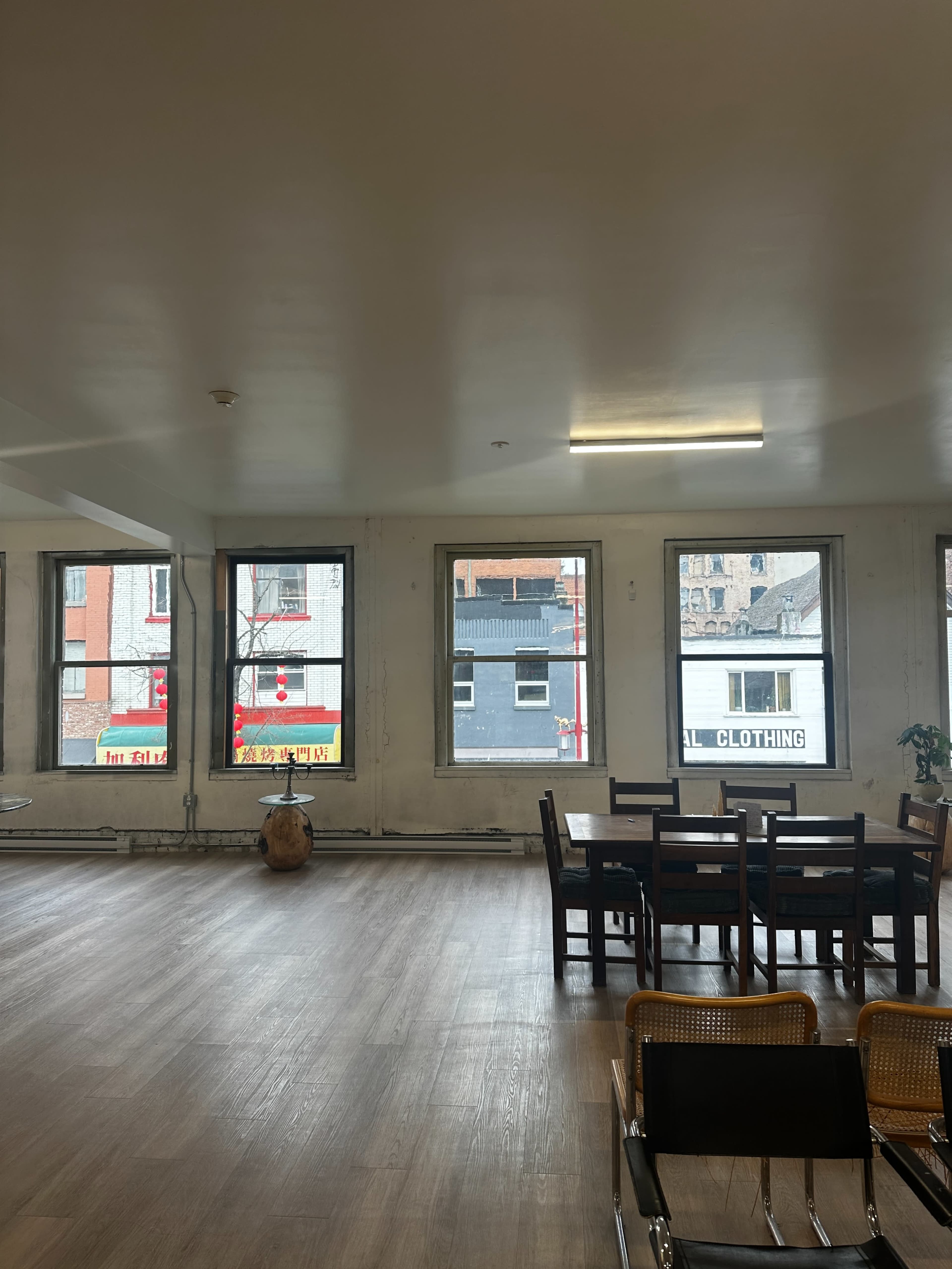 A spacious, empty room with wooden flooring, several large windows, and a dining table with chairs.