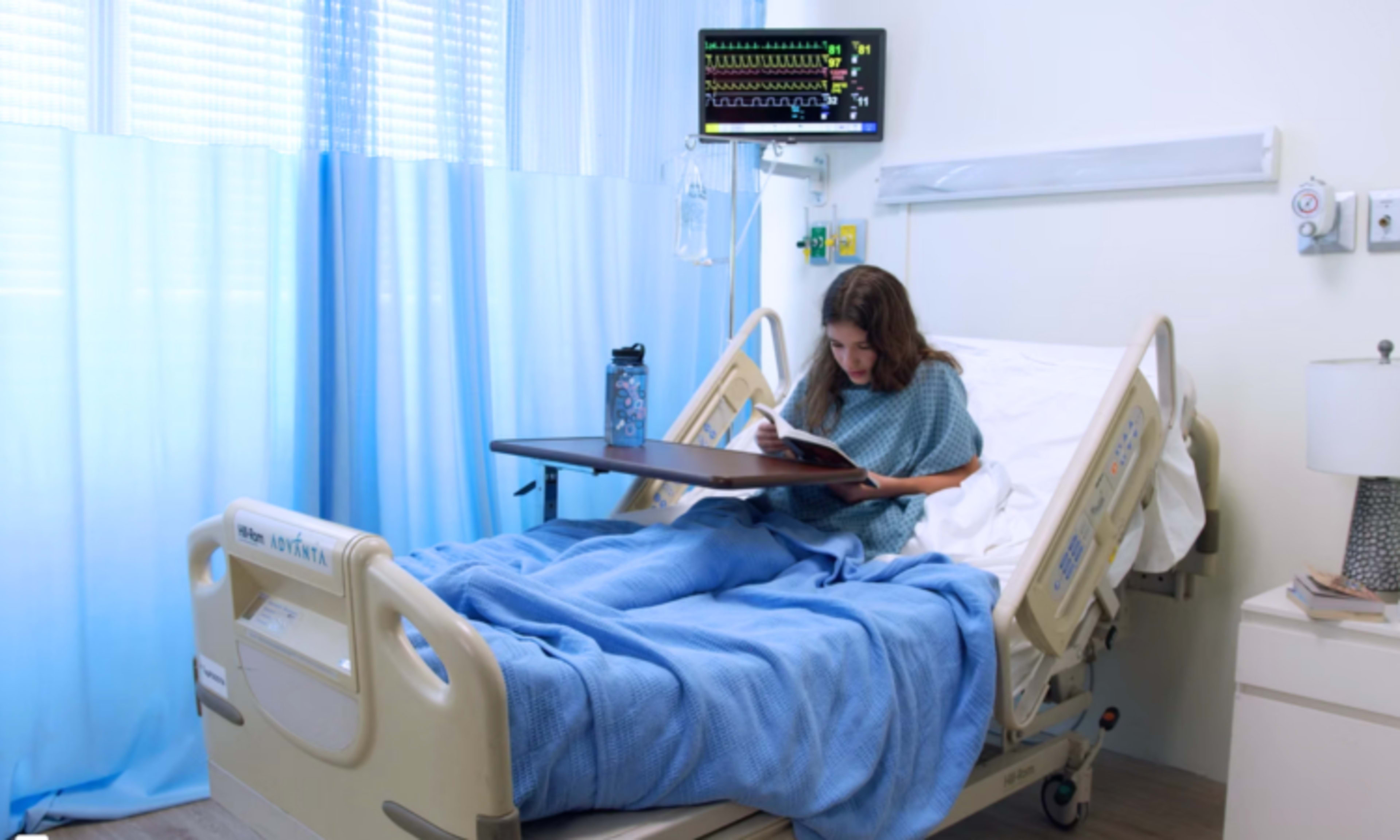 A young patient sits on a hospital bed, reading a book in a brightly lit hospital room equipped with medical monitoring equipment.