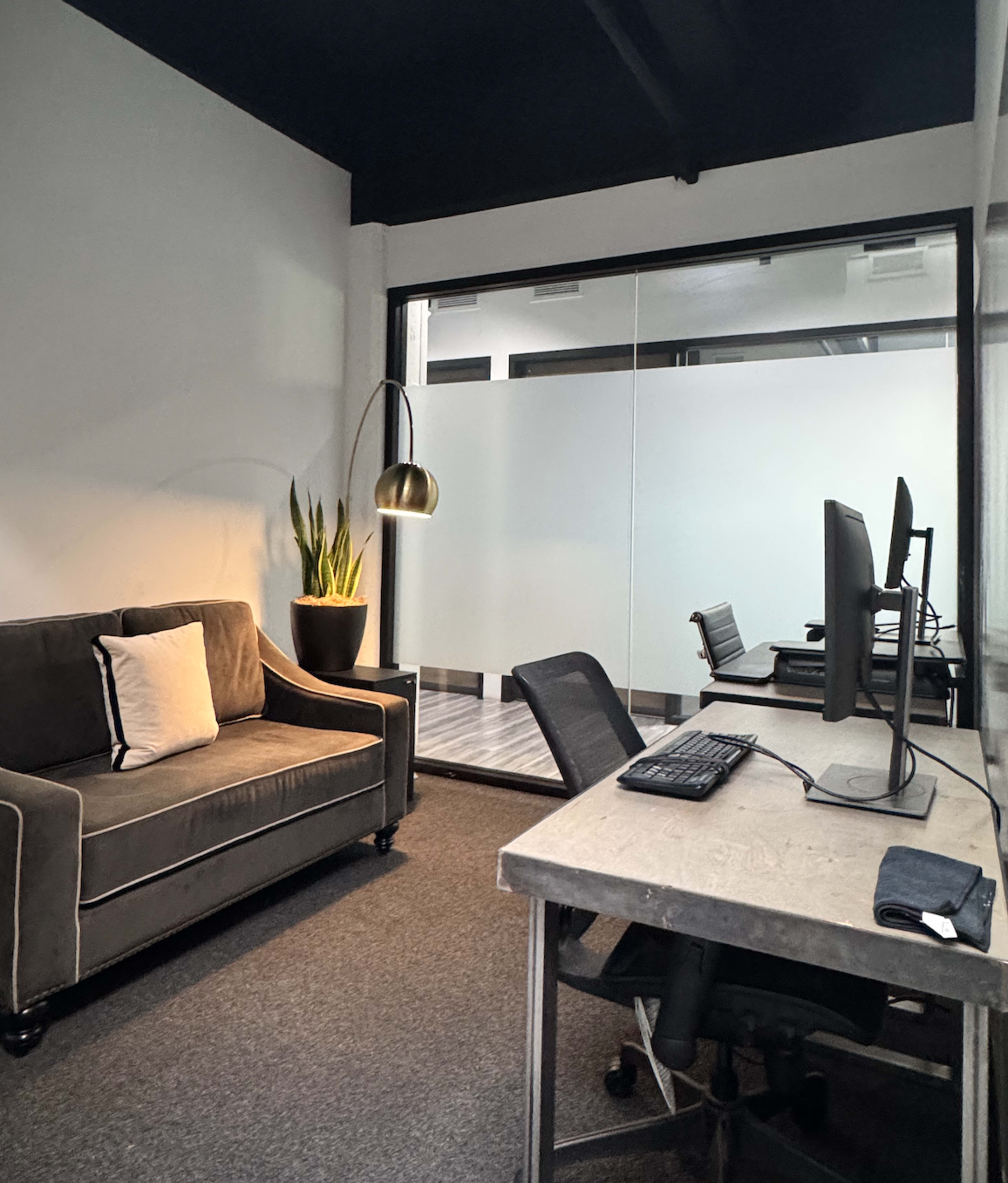 The image shows a modern office space with a sofa, a potted plant, and two desk setups with computers.