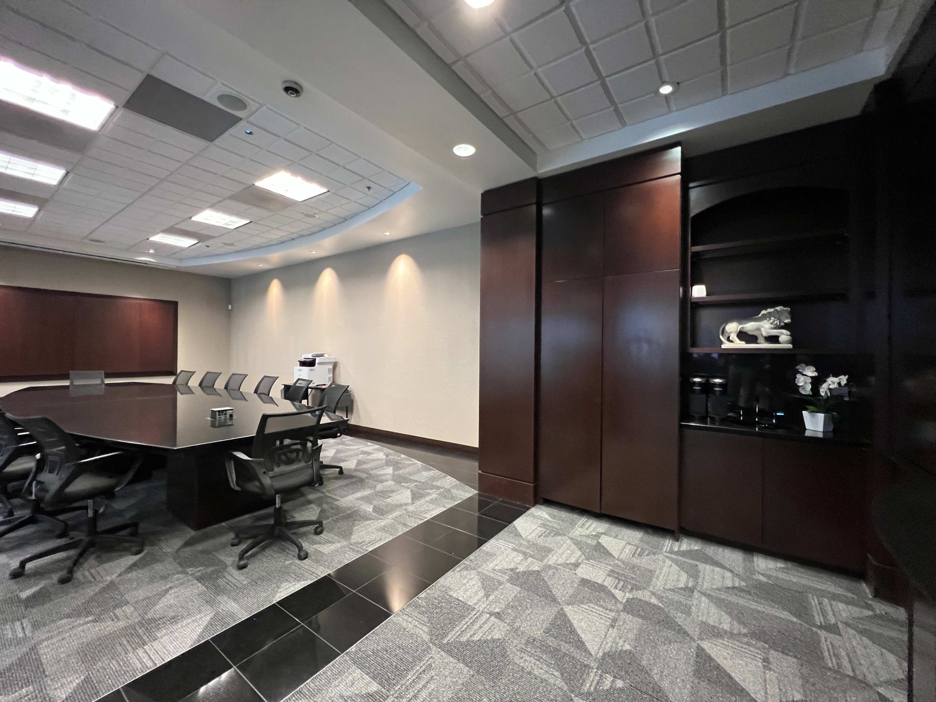 A modern conference room features a large rectangular table surrounded by black chairs, with dark wood paneling and a sleek corner cabinet displaying decor.