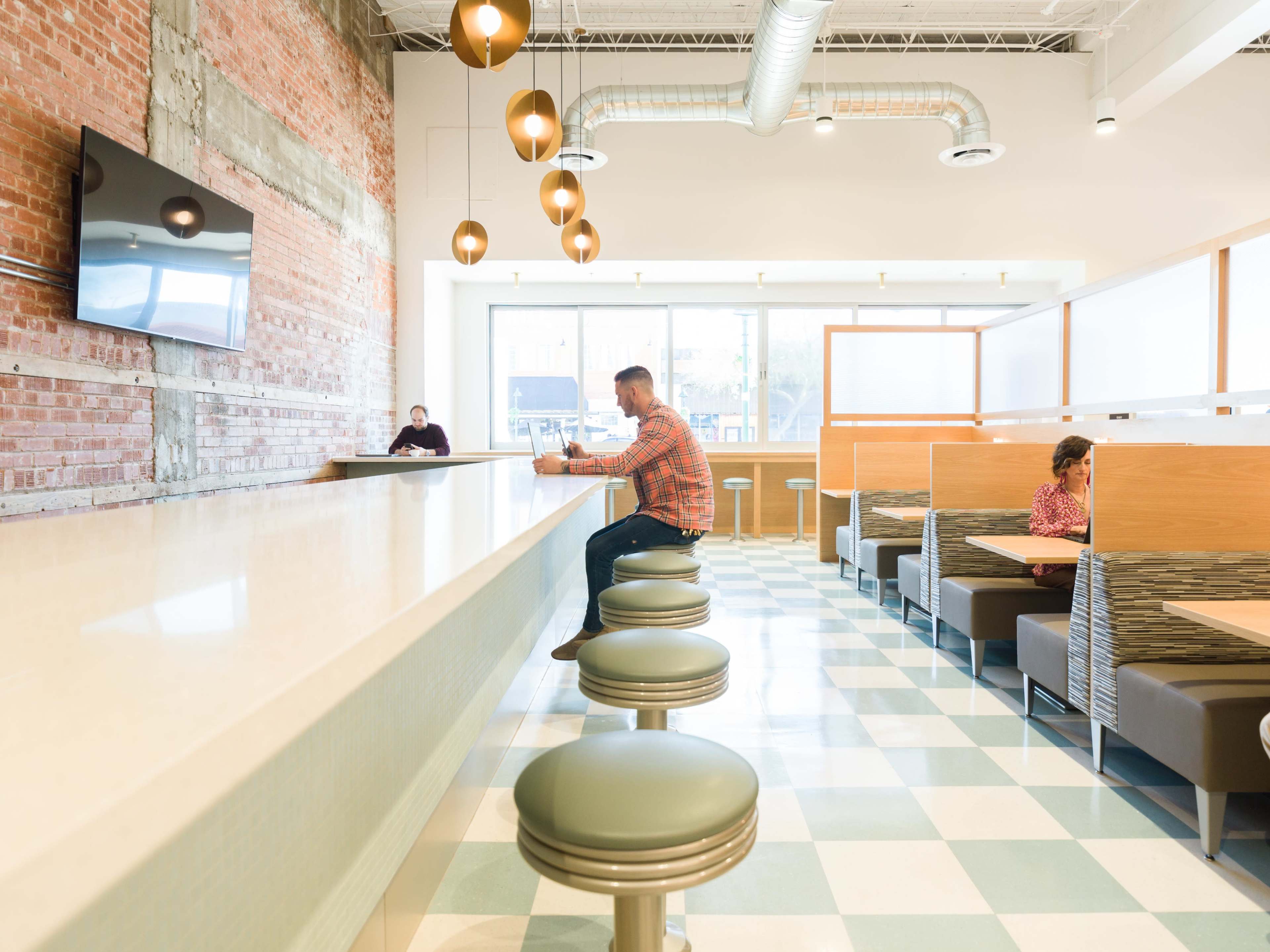 The image shows a modern cafe interior with a long counter, stools, and wooden booths, featuring a mix of exposed brick and clean lines.