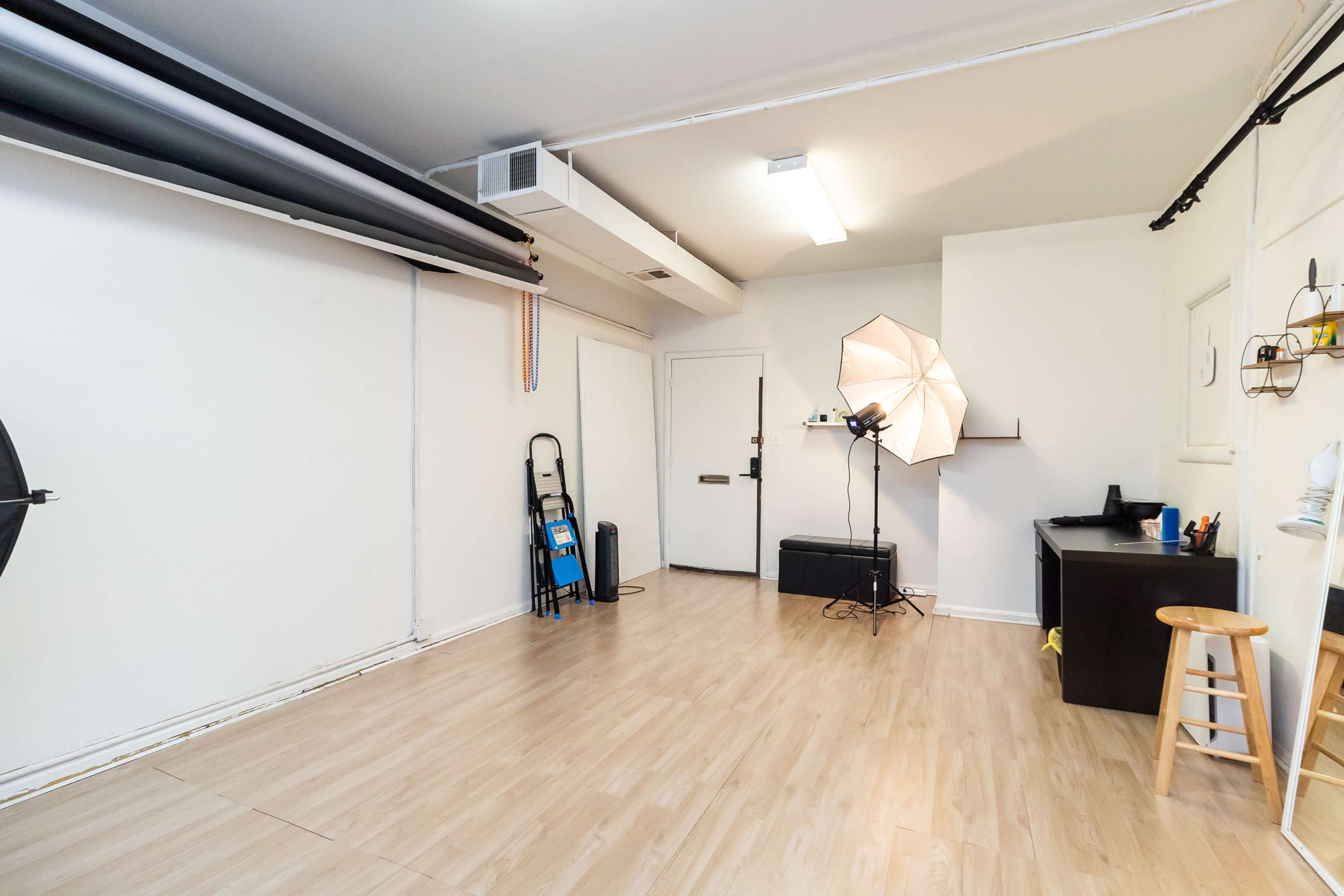The image shows a studio space with a light stand, a black stool, a door, and various equipment against the walls, all on a wooden floor.