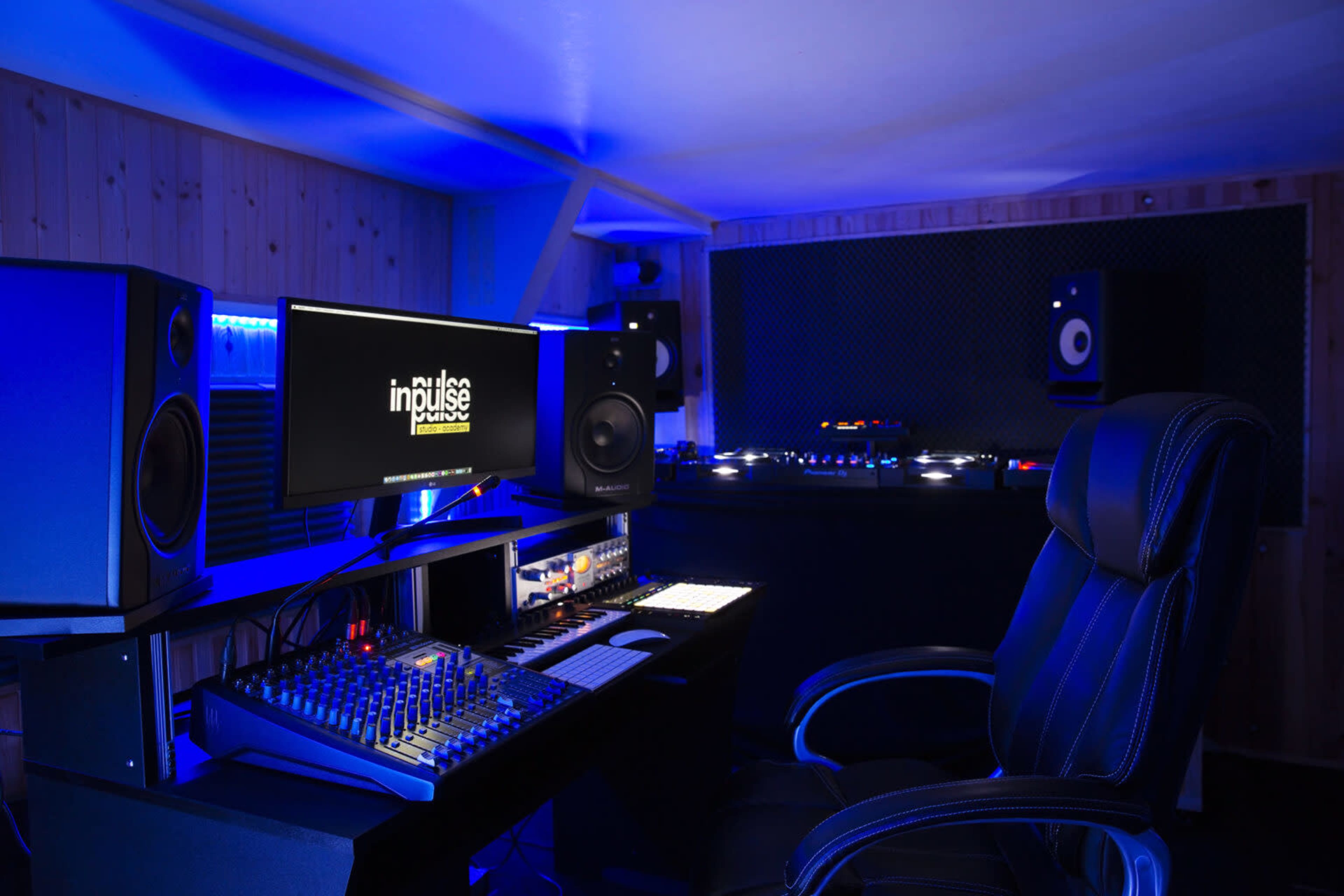 A recording studio features a central mixing console, large speakers, and a computer monitor displaying the logo "impulse," all illuminated by blue lighting.