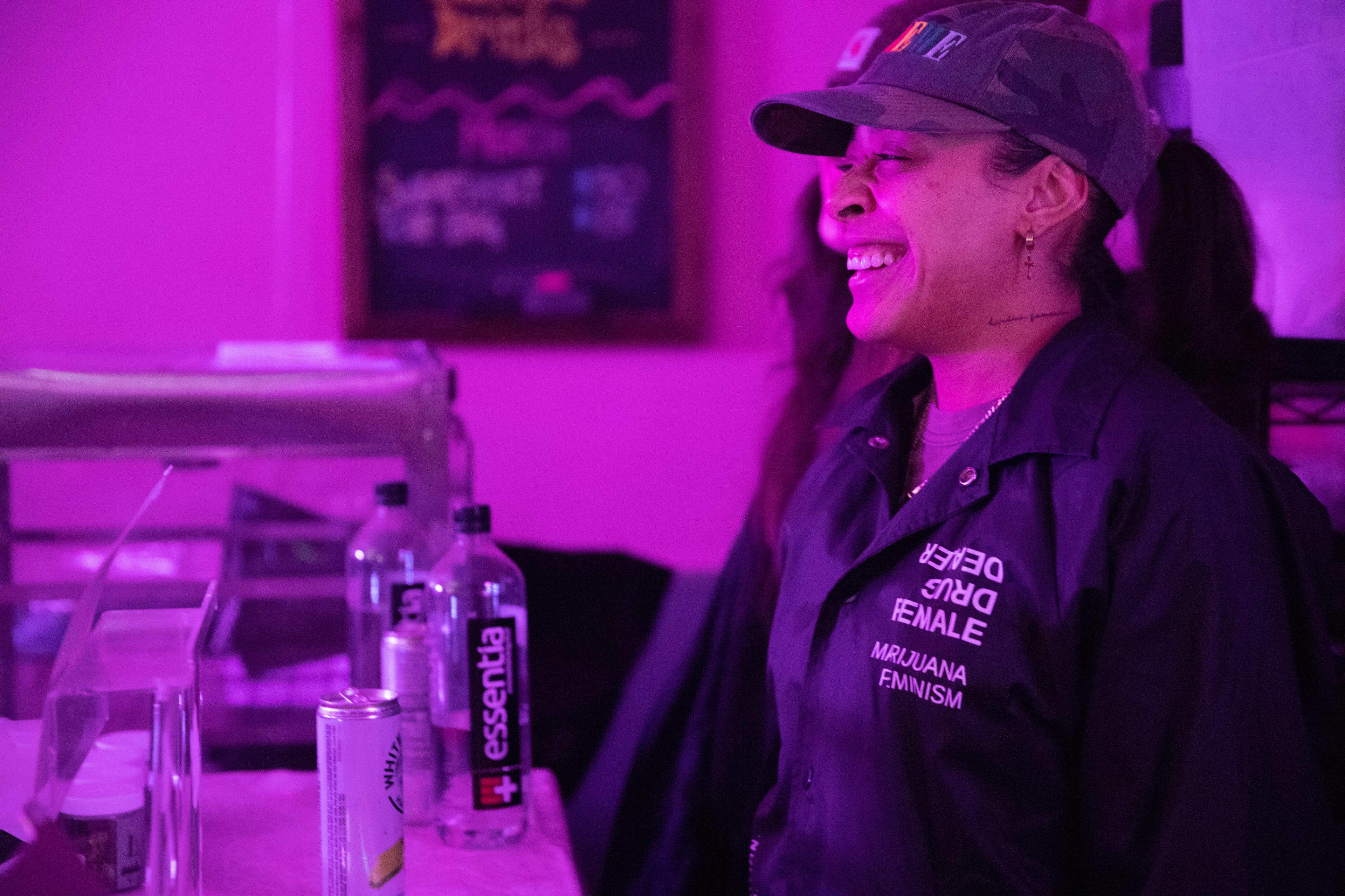 A woman wearing a dark jacket and a cap stands behind a counter, smiling in a brightly lit, purple-toned space.