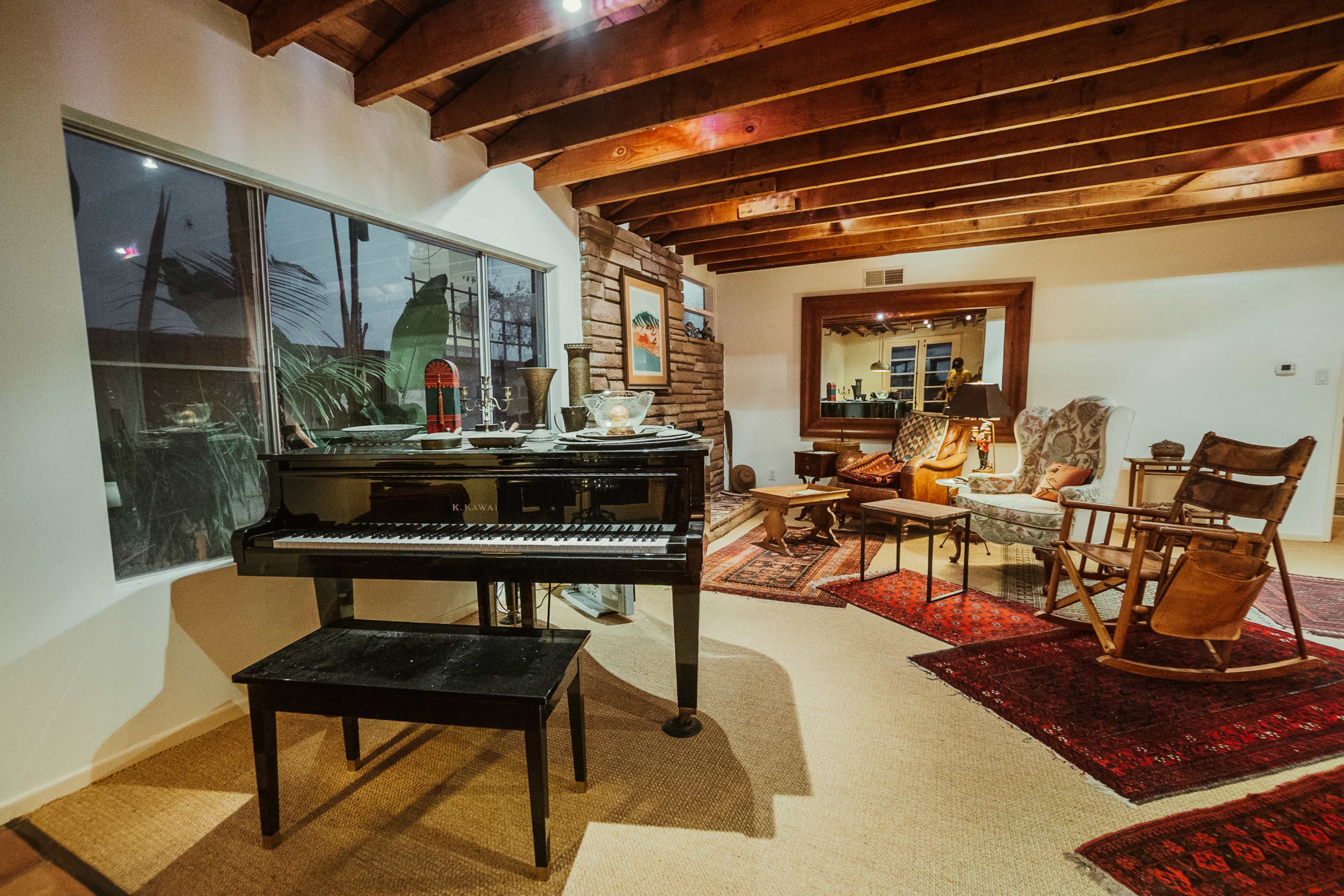 A black piano is positioned next to a window in a cozy living room featuring wooden beams, a rocking chair, and various decorative rugs.
