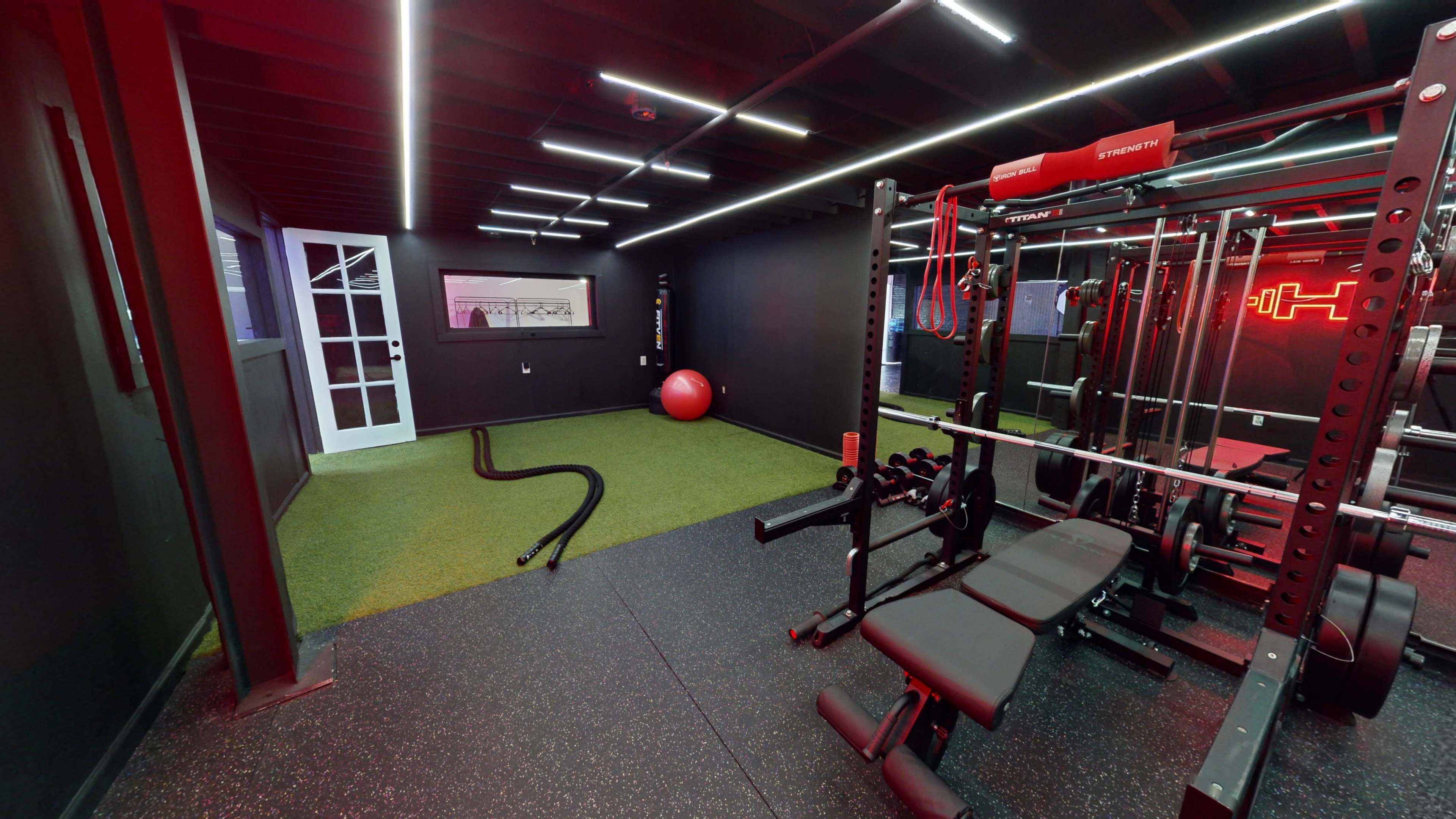 A modern gym interior featuring exercise equipment, a large red exercise ball, and artificial grass flooring under ambient lighting.