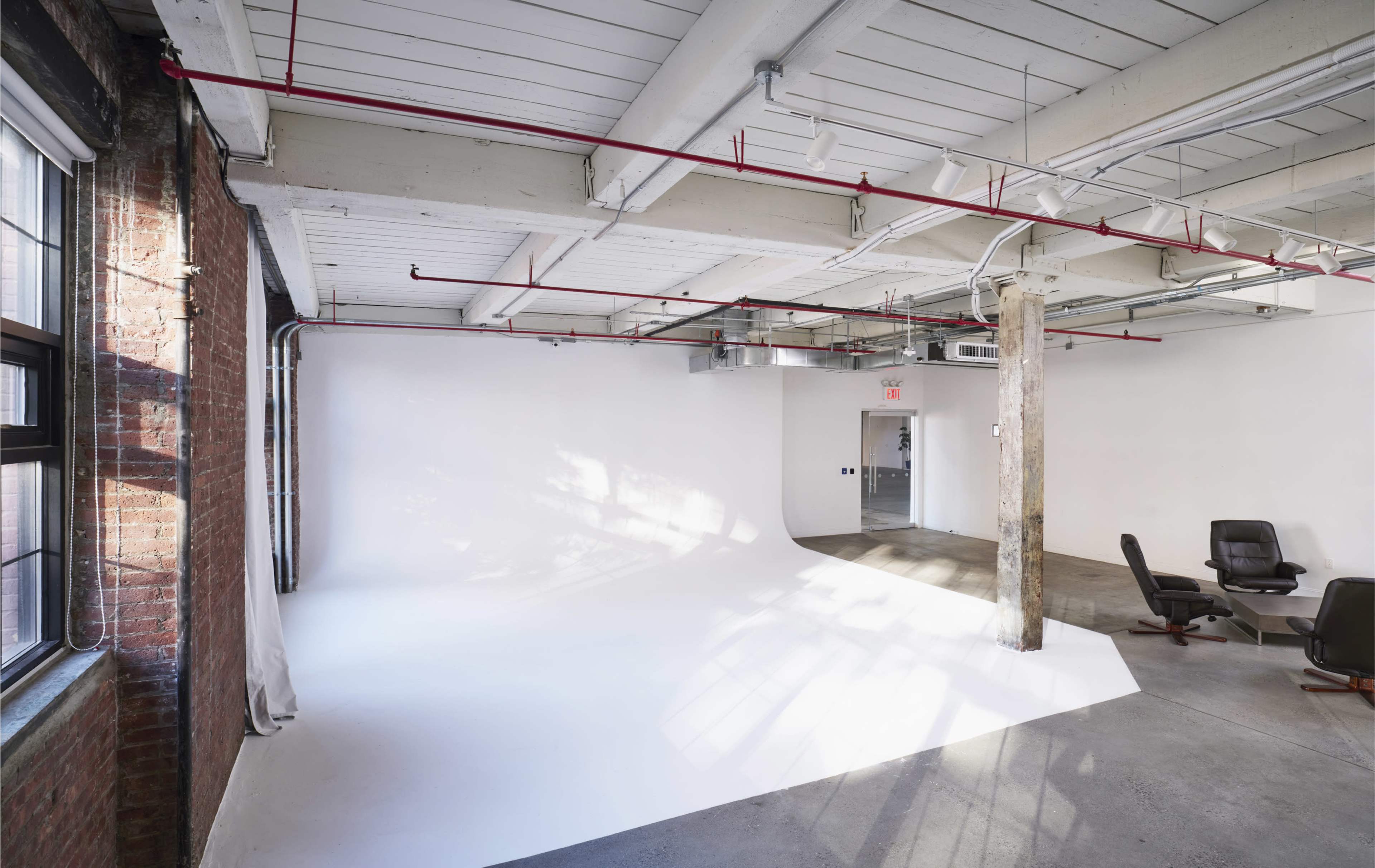 The image shows a spacious studio with exposed brick walls, a smooth white backdrop, and industrial-style features including overhead pipes and a support column.