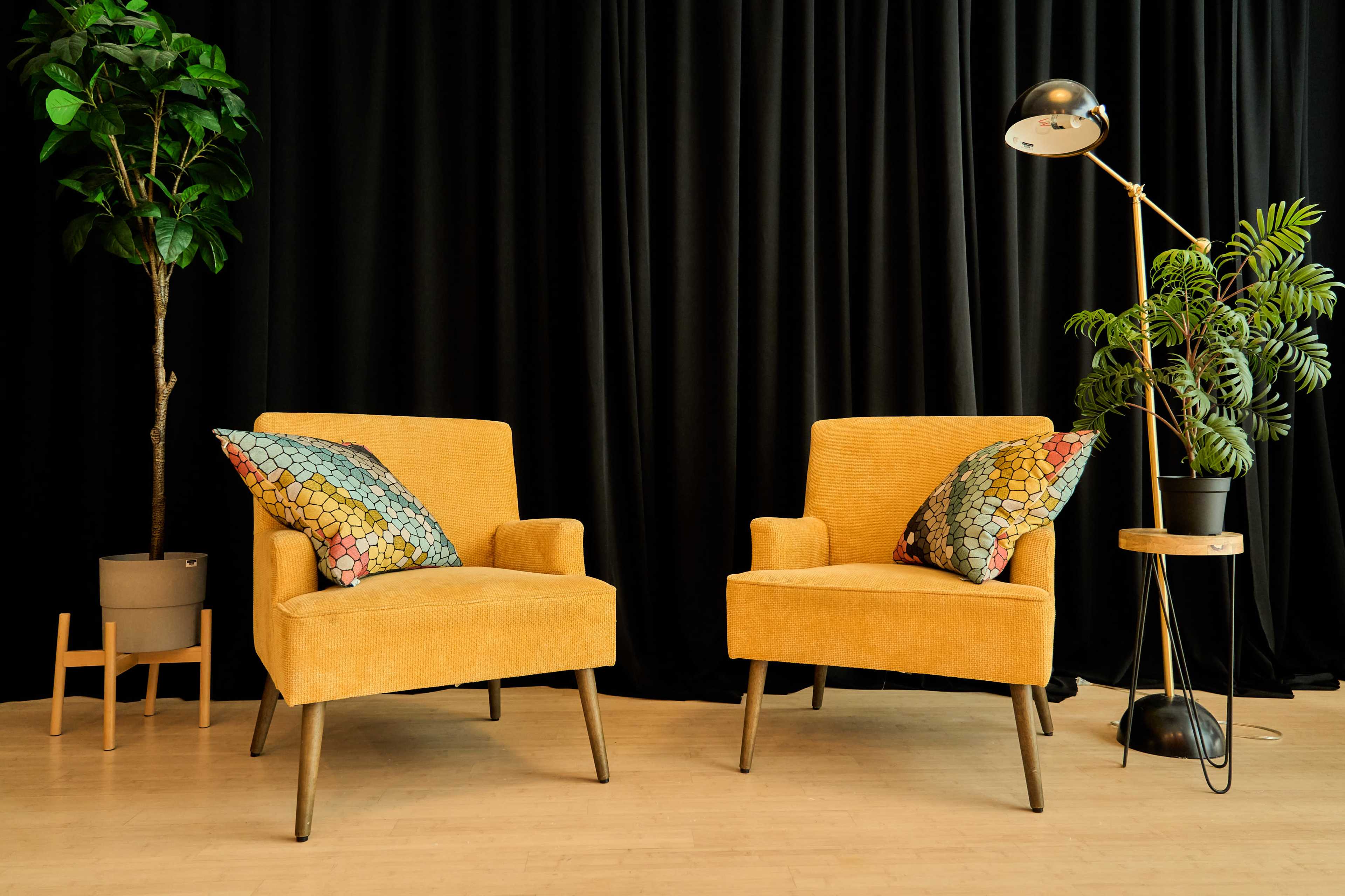 The image features two yellow chairs with patterned pillows, a potted plant beside each chair, and a black drape in the background.