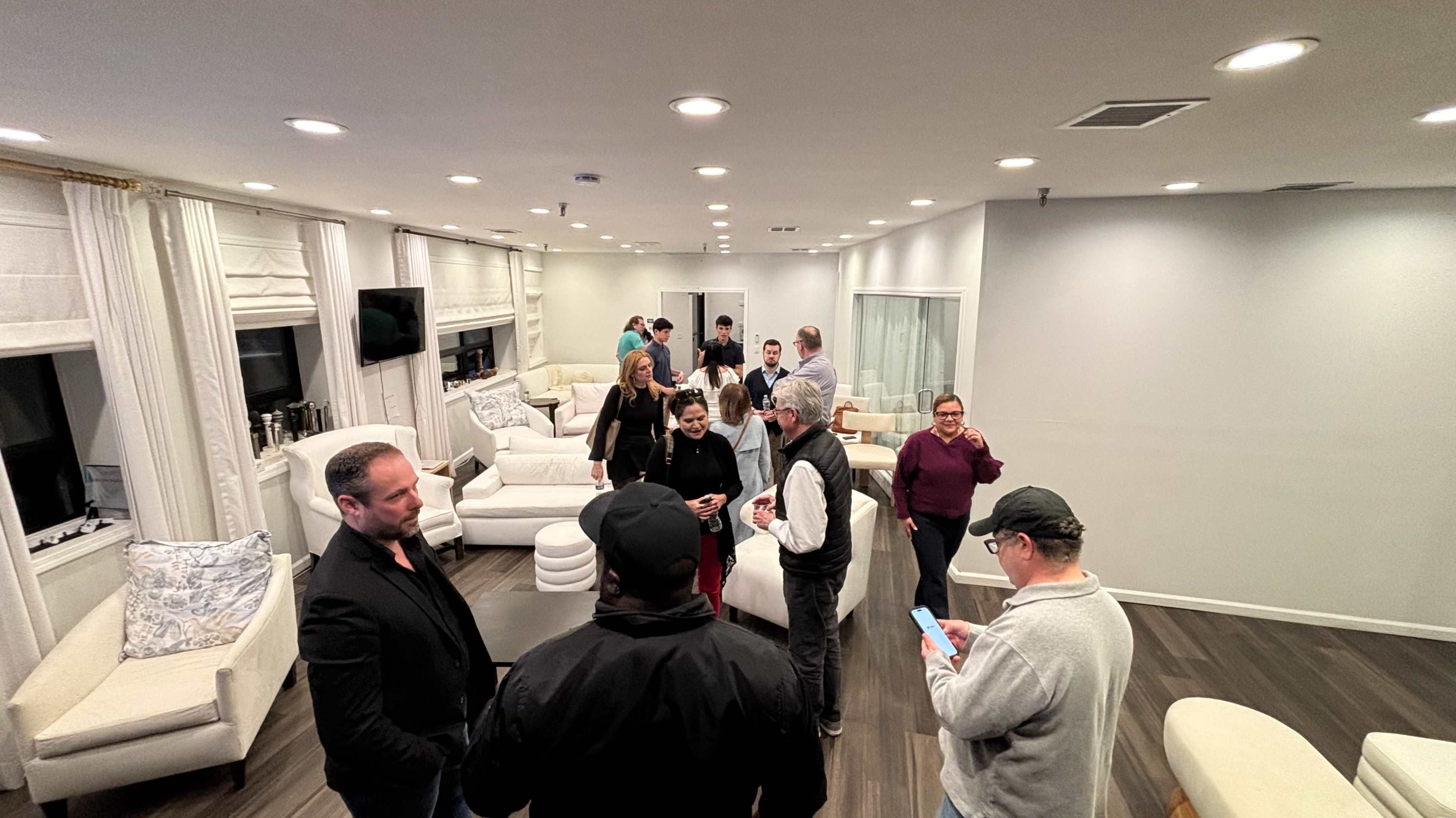 A group of people socialize in a well-lit, modern lounge with white furniture and large windows.