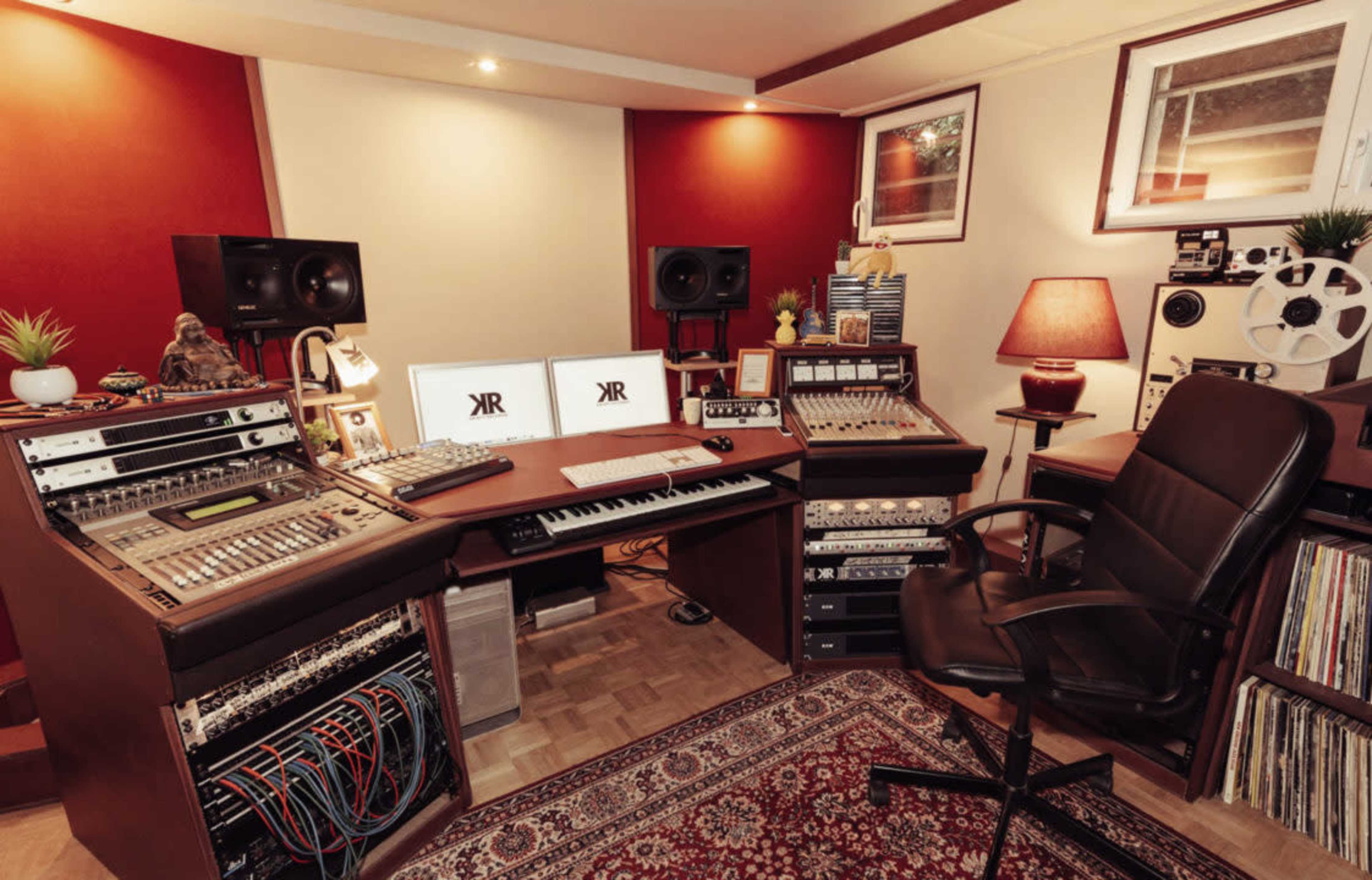 A music studio with a desk equipped with audio equipment, two computer monitors, a chair, and various recording devices against a backdrop of red and neutral walls.