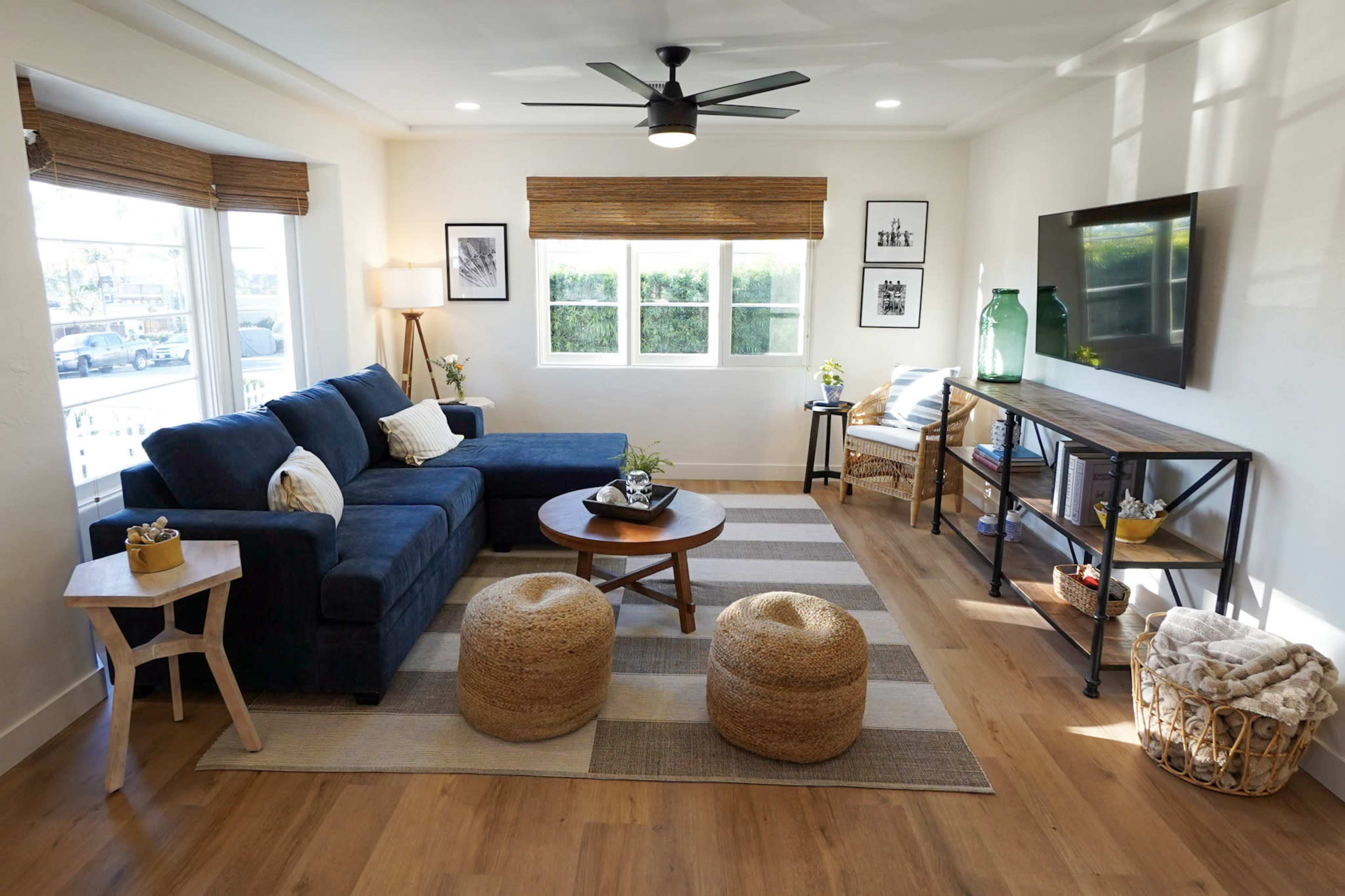 The living room features a blue sofa, a round wooden coffee table, two woven poufs, and a side table, all arranged around a patterned rug under natural light from large windows.