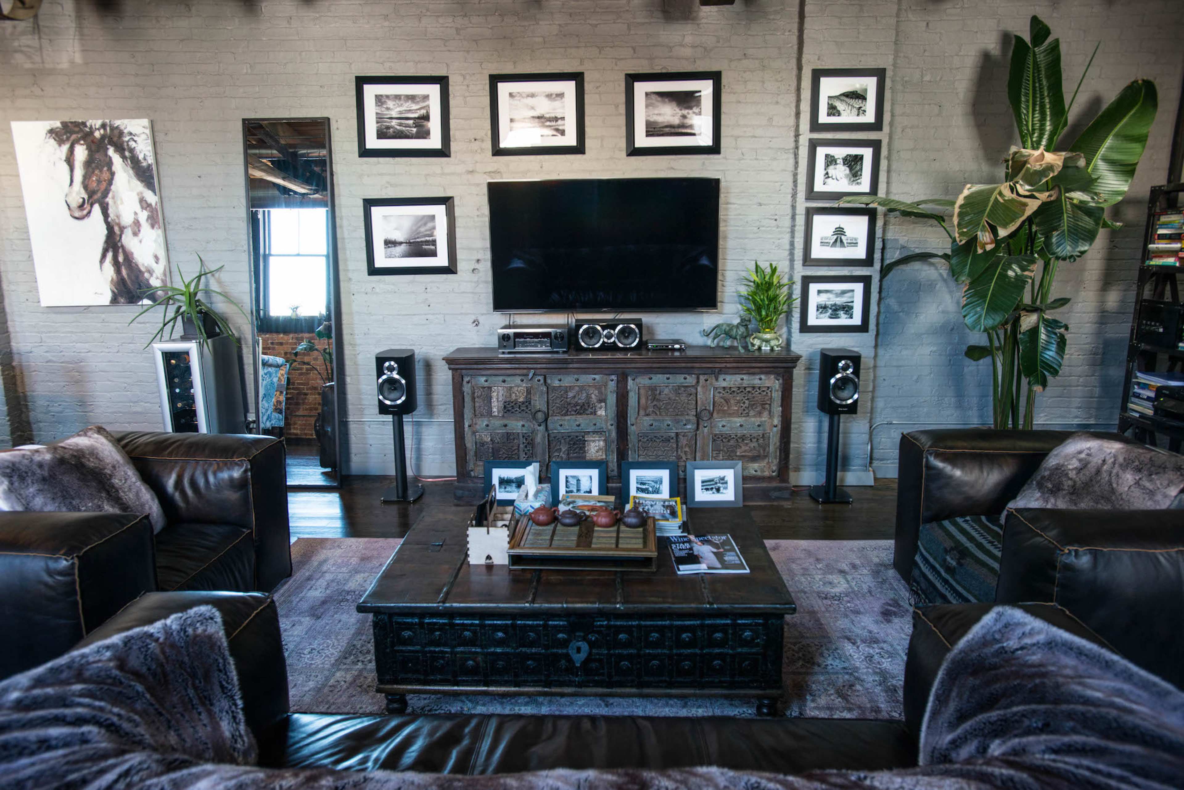 A living room features a large television mounted on a brick wall, flanked by framed photographs, with leather sofas surrounding a coffee table and a decorative plant in the corner.
