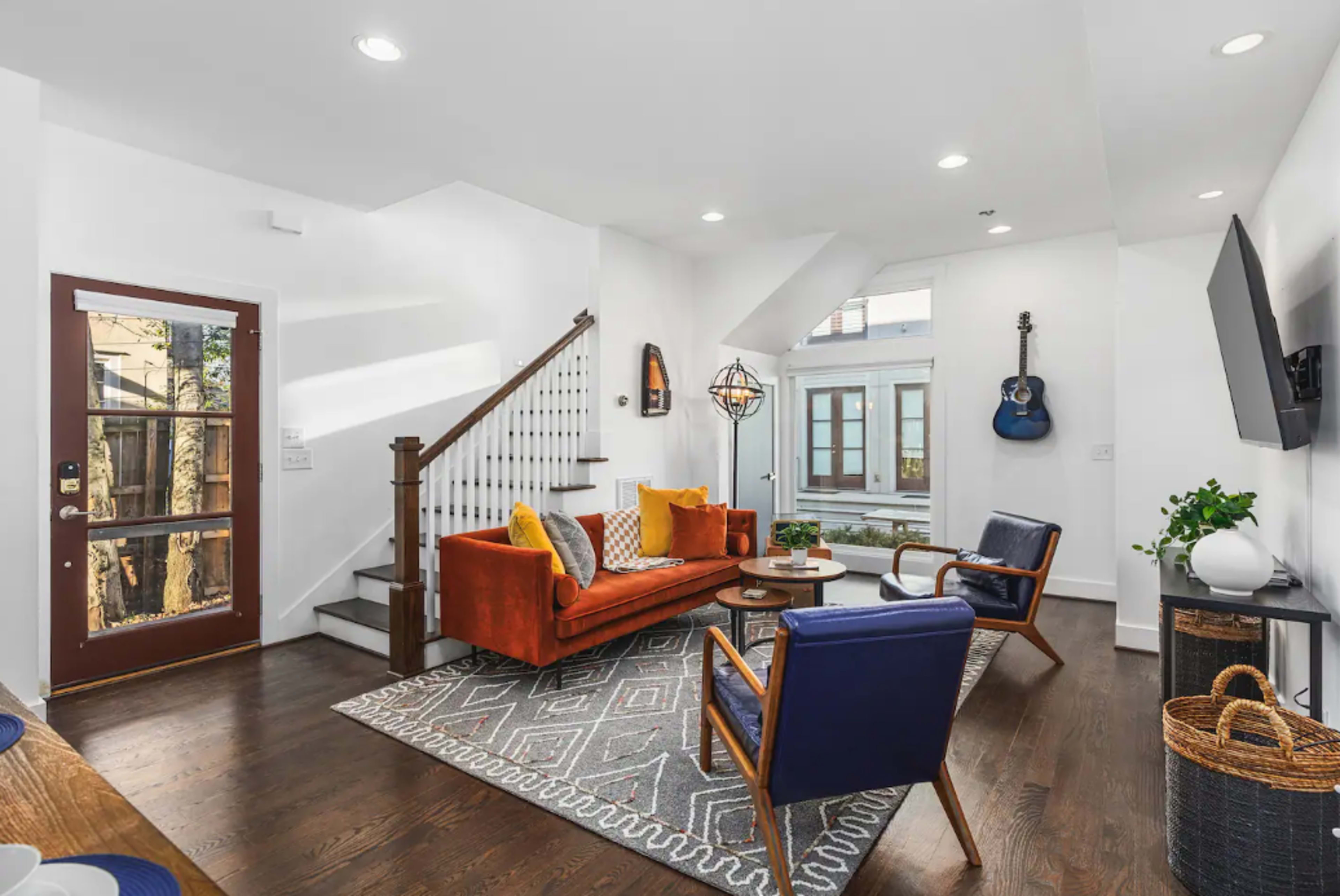 A modern living room featuring a red sofa, blue armchairs, a patterned area rug, and a staircase leading to an upper level.