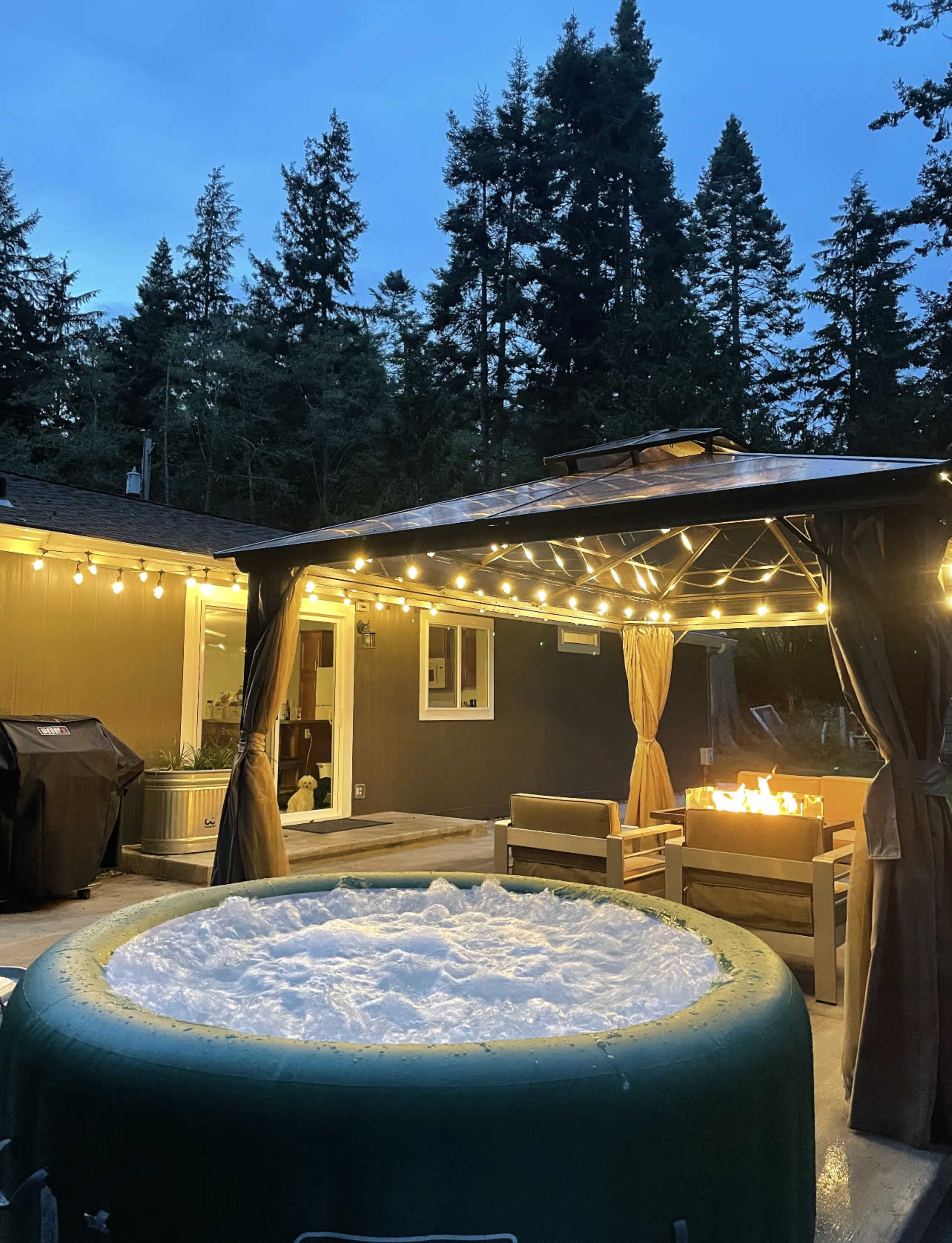 A hot tub surrounded by outdoor seating and a fire pit, illuminated by string lights against a wooded backdrop at dusk.