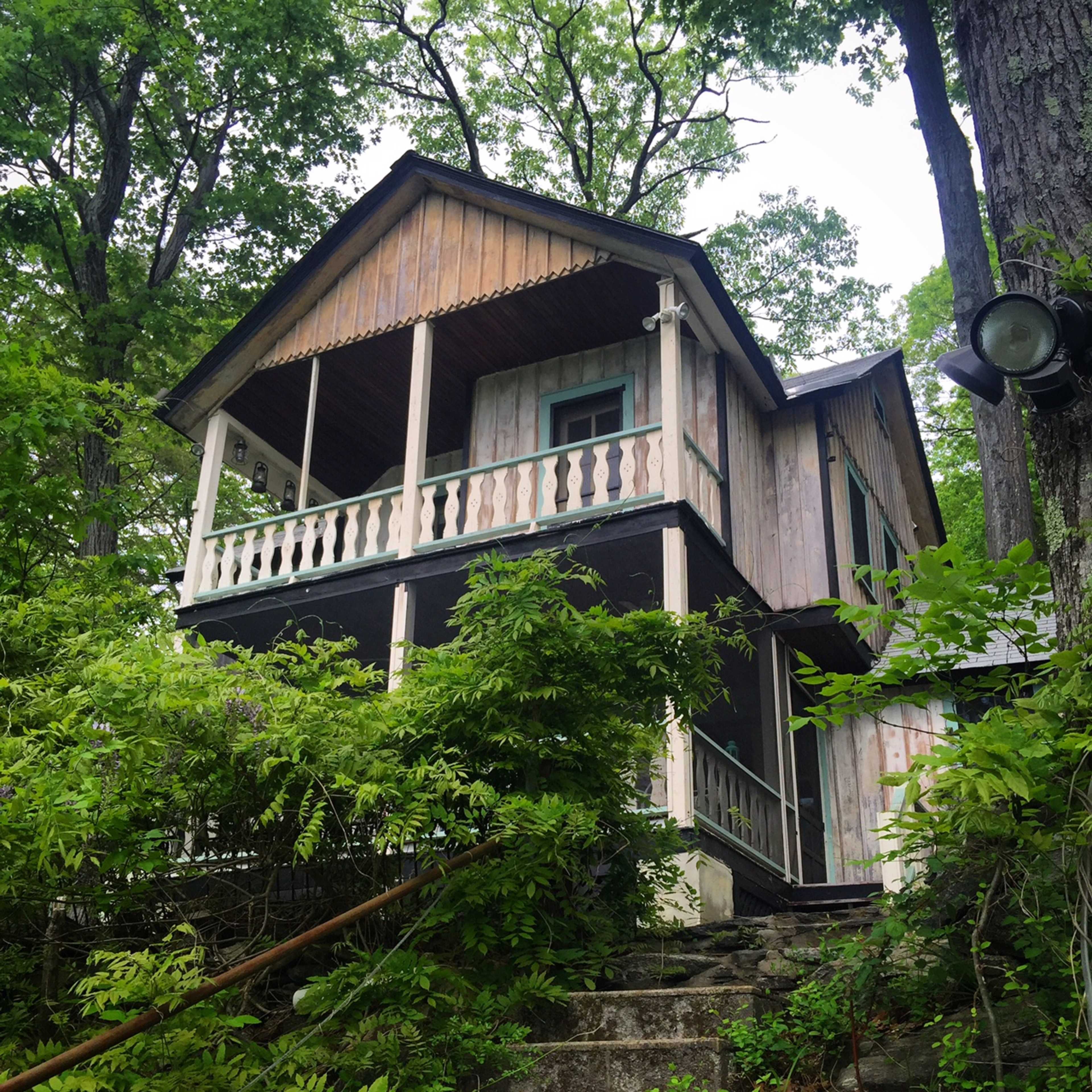 A wooden house with a balcony is situated among dense greenery on a hillside.