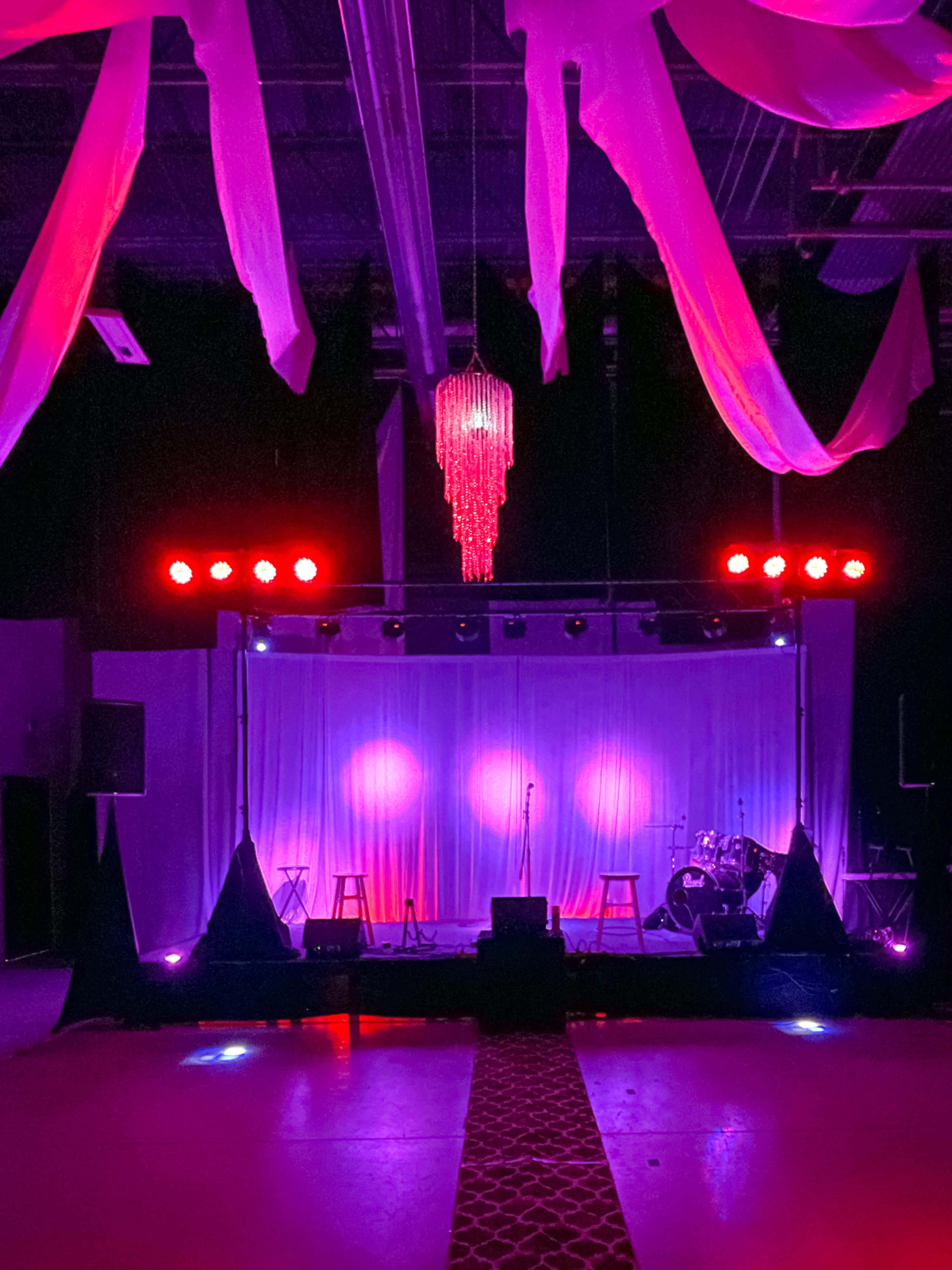 A performance stage is set up with a microphone, stools, and musical instruments under colorful lighting and draped fabric overhead.
