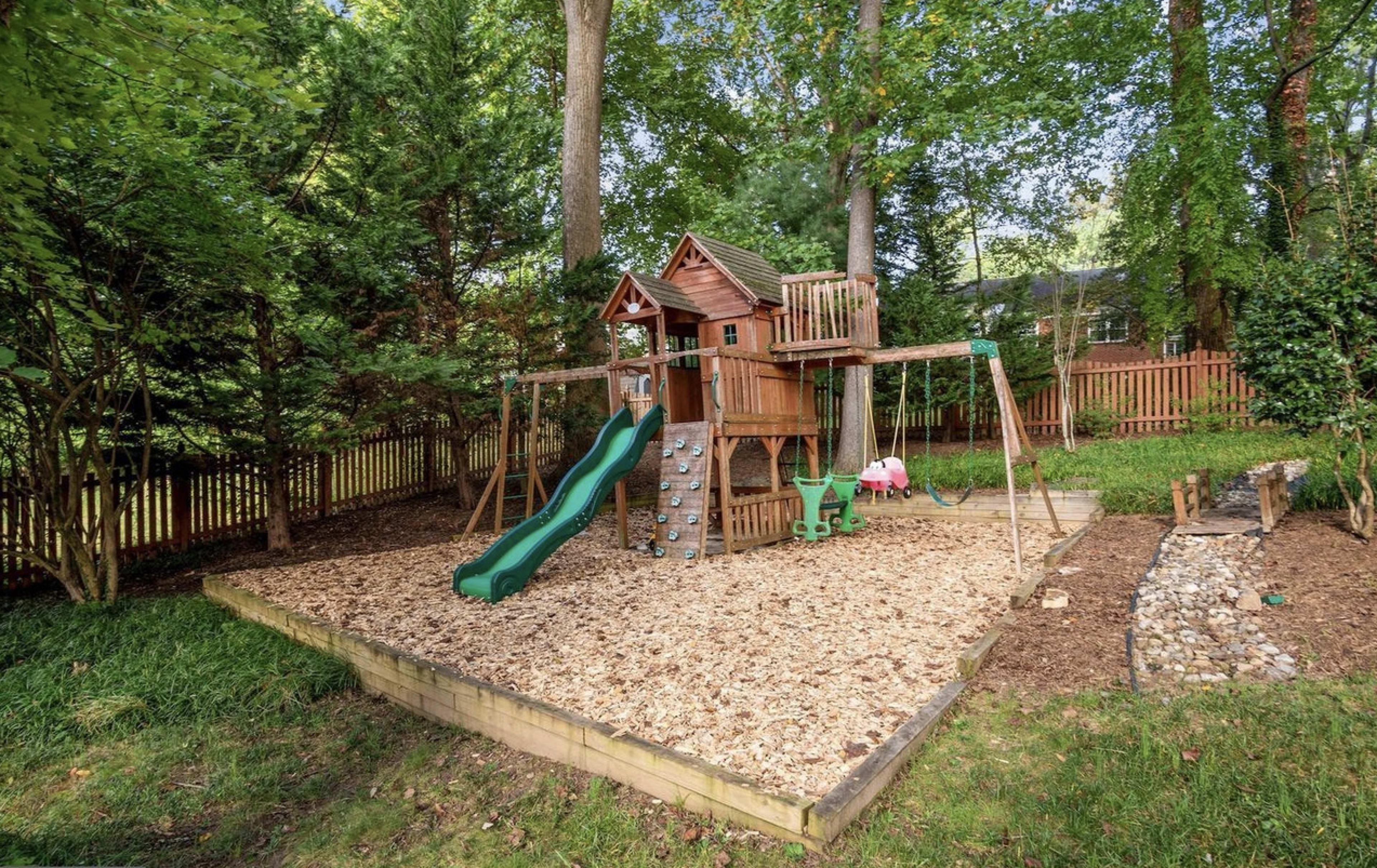 A wooden playset with slides and climbing features stands on a mulch-covered area surrounded by trees and a wooden fence.