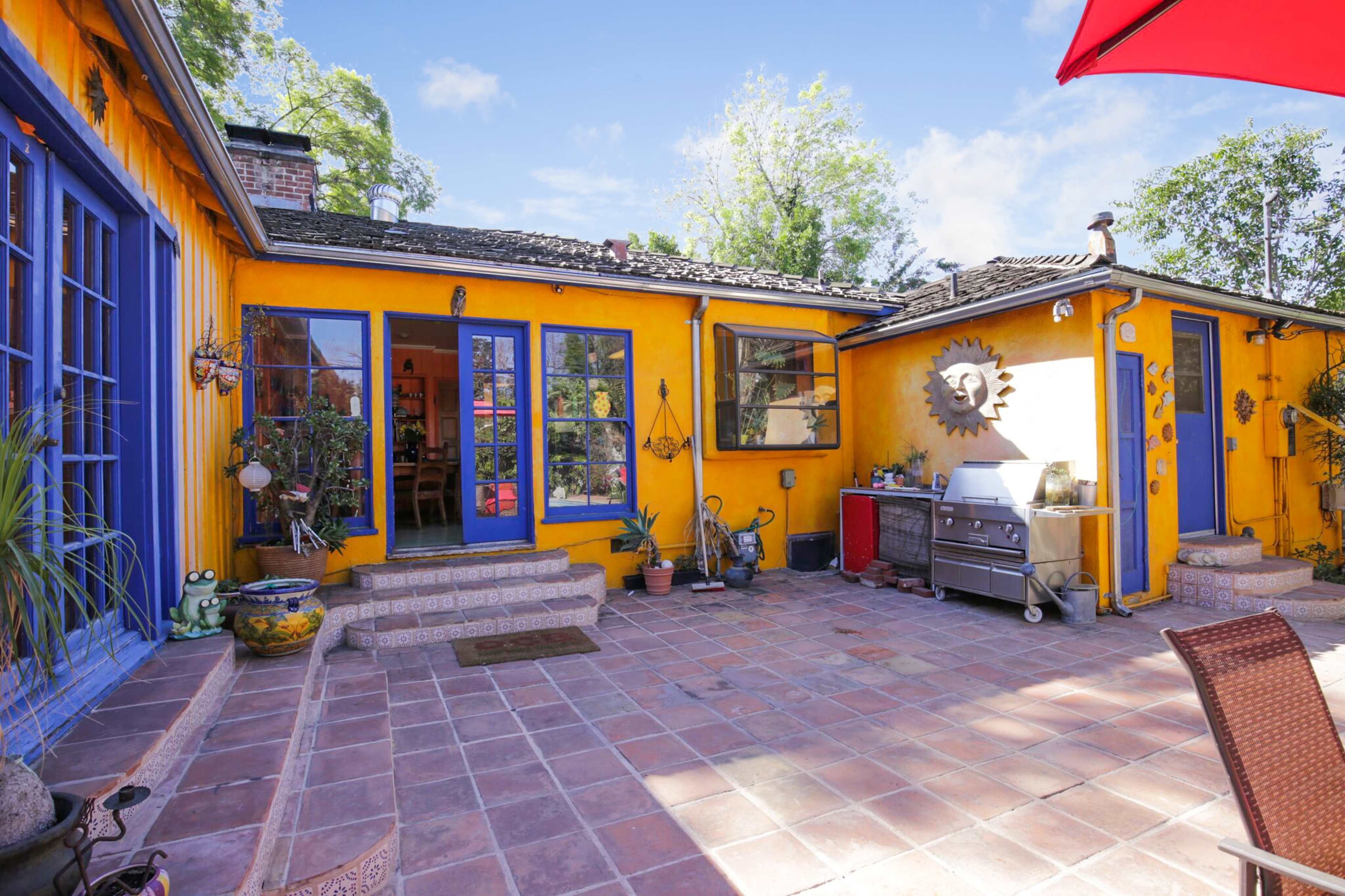 The image shows a vibrant courtyard with yellow walls, blue doors and windows, tiled flooring, and a BBQ grill.