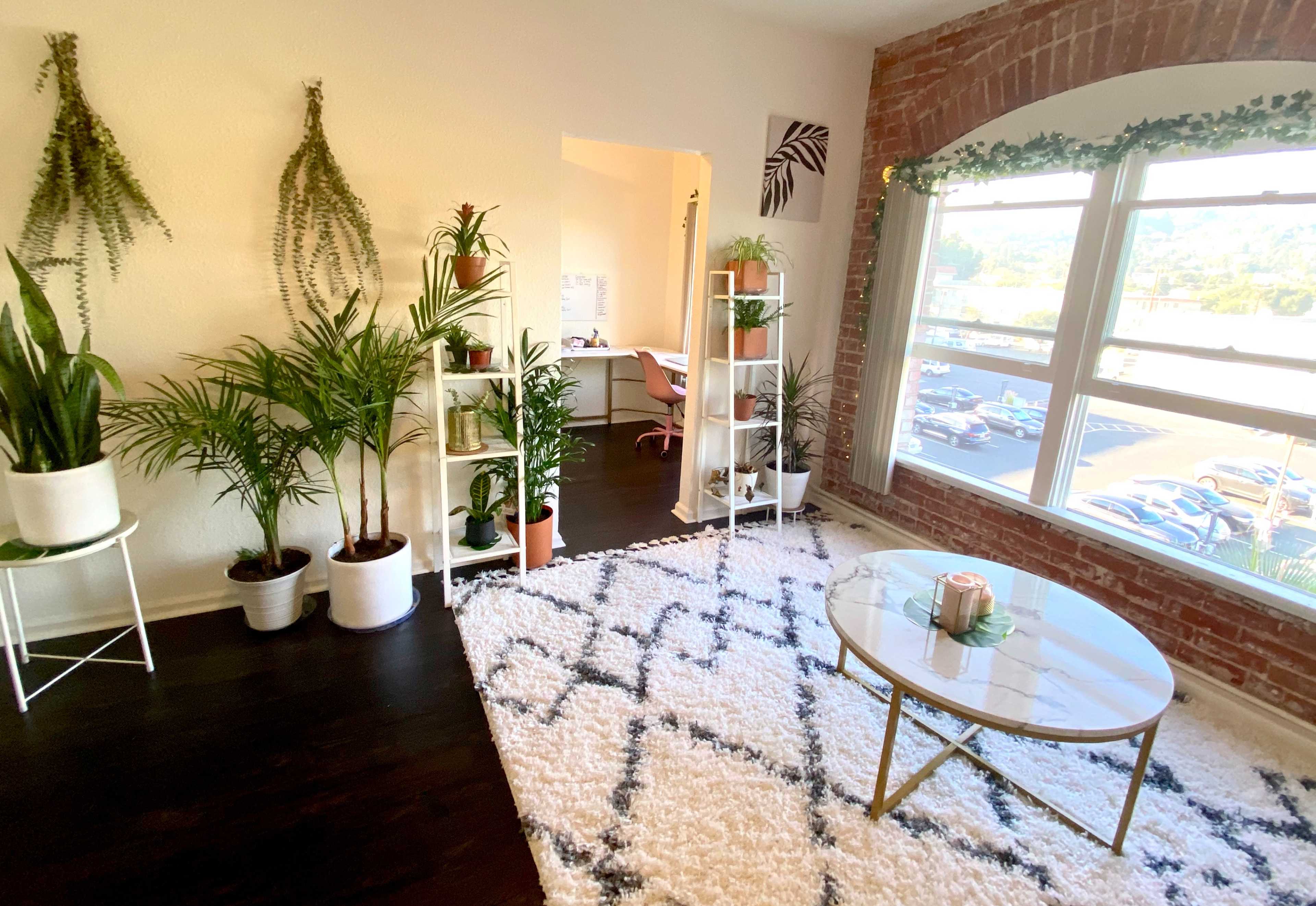 A bright, modern living space featuring a white rug, a round coffee table, various indoor plants, and large windows with views of parked cars outside.