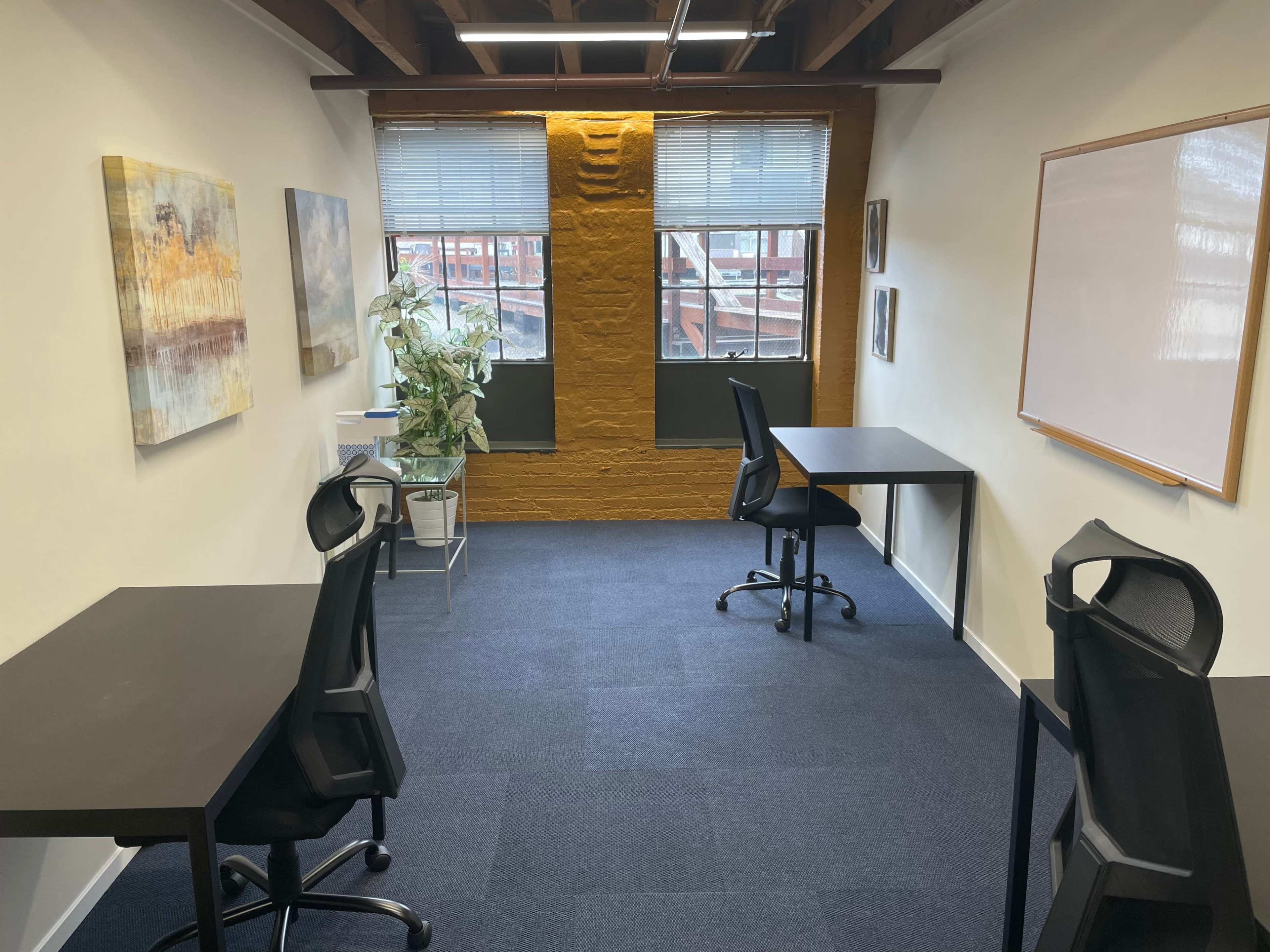 The image shows a modern office space with two black desks and chairs, large windows, and a plant, set against a textured yellow wall.
