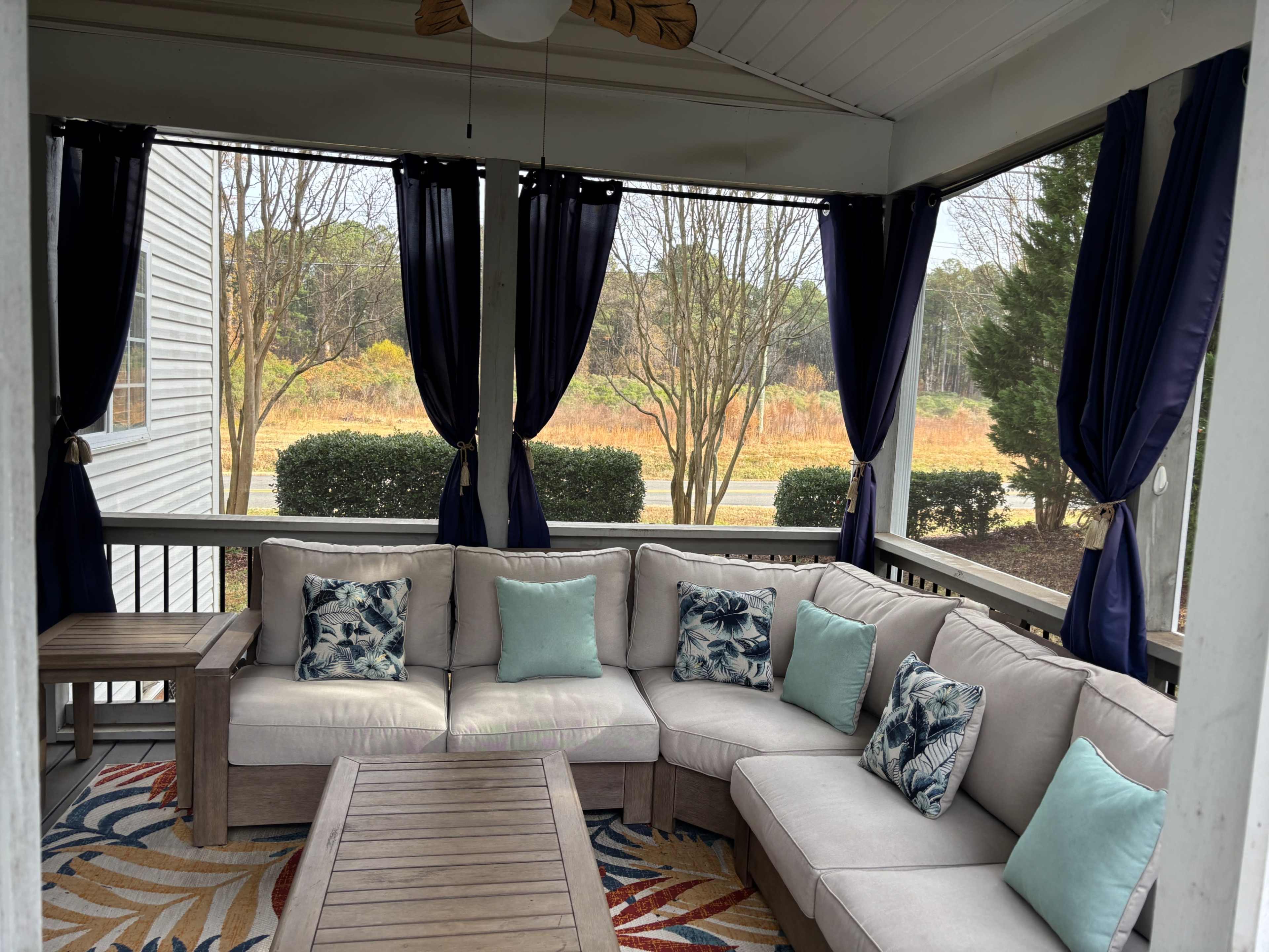 The image shows a screened porch with a light-colored sectional sofa adorned with turquoise and floral cushions, alongside a wooden coffee table, and windows draped with dark blue curtains.
