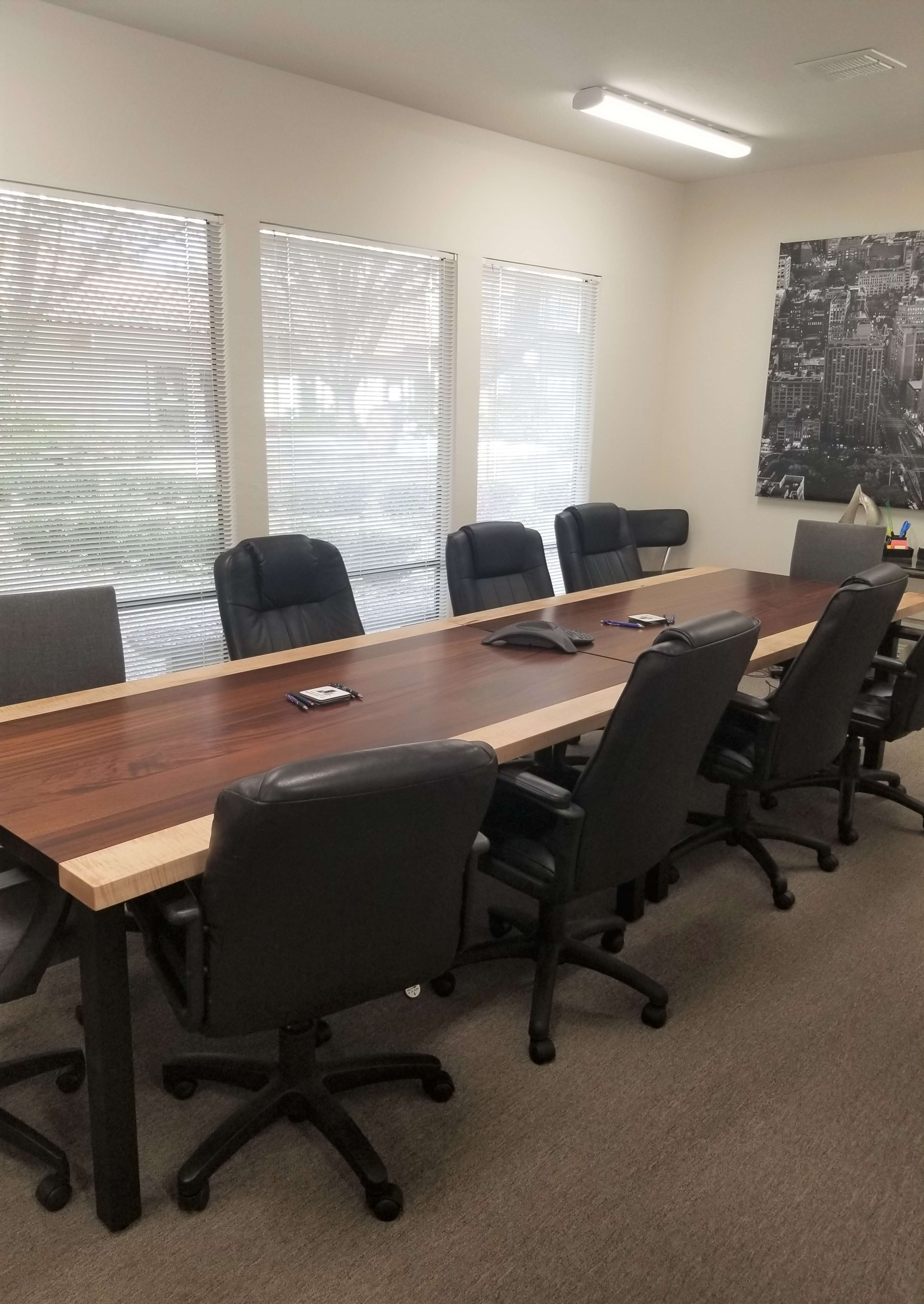 A conference room features a large wooden table surrounded by black rolling chairs, with windows letting in natural light.