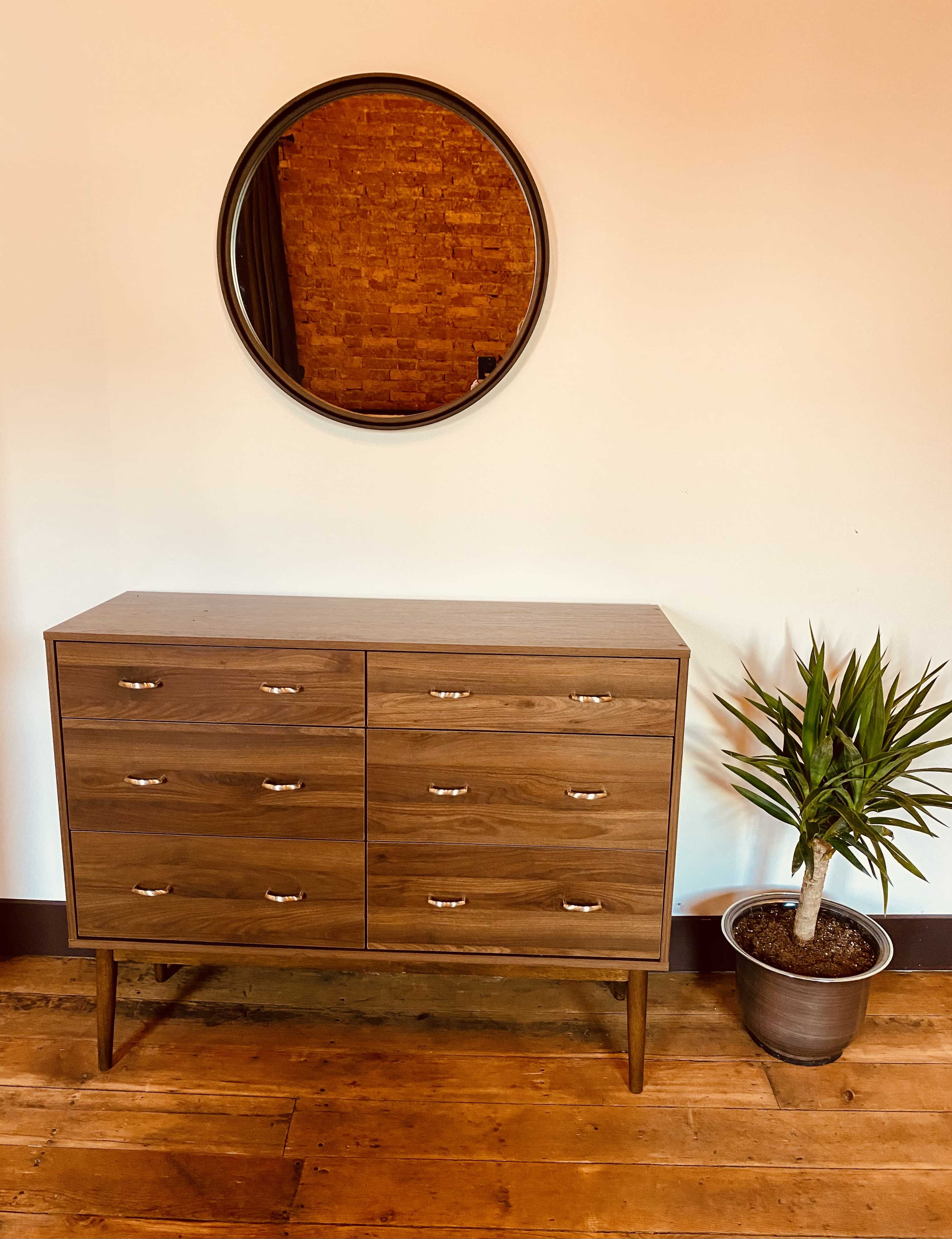 A wooden dresser with six drawers is positioned beneath a round mirror, accompanied by a potted plant in the corner of the room.