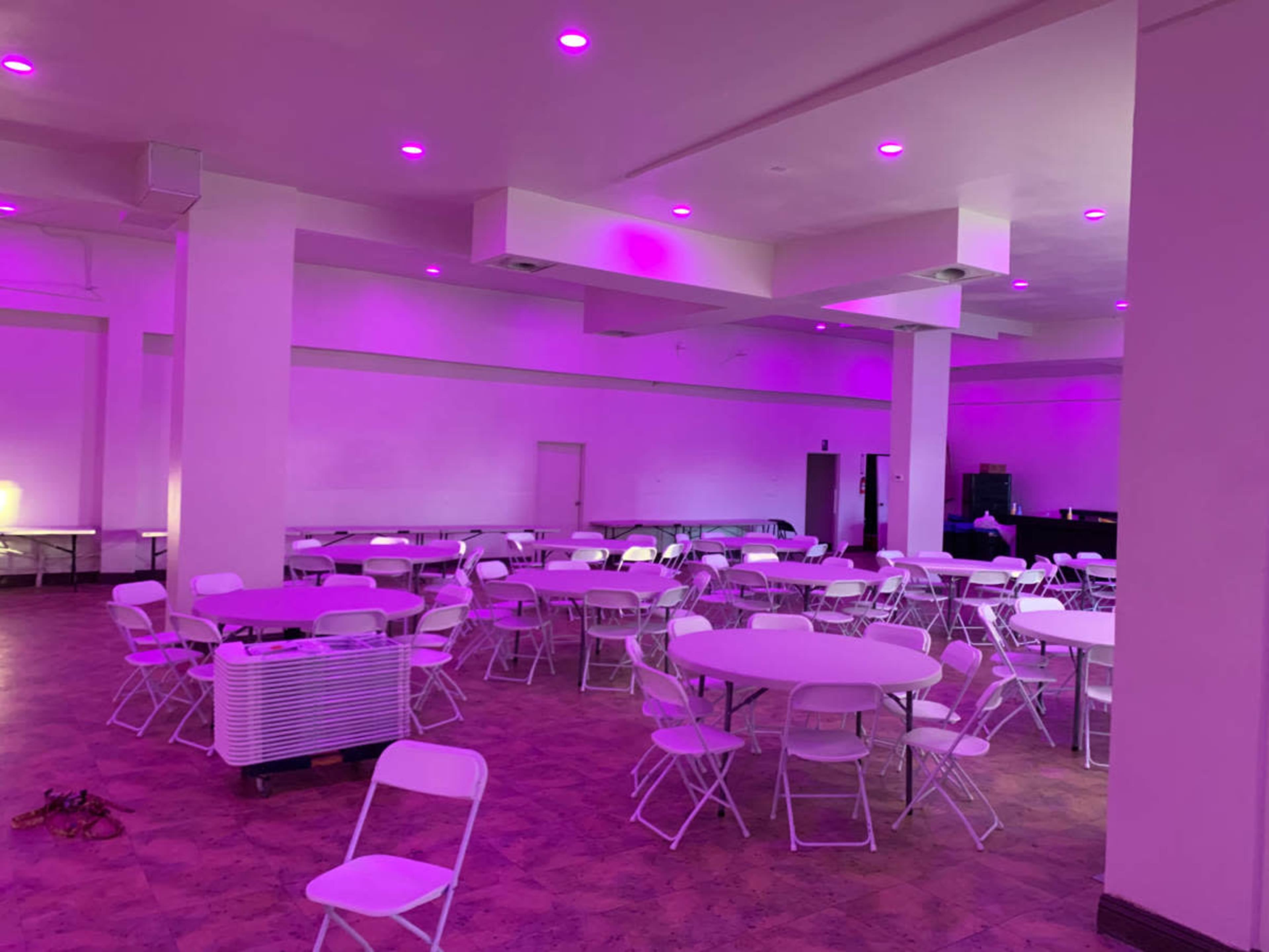 A spacious indoor area features several round tables surrounded by white chairs, illuminated by purple ceiling lights.
