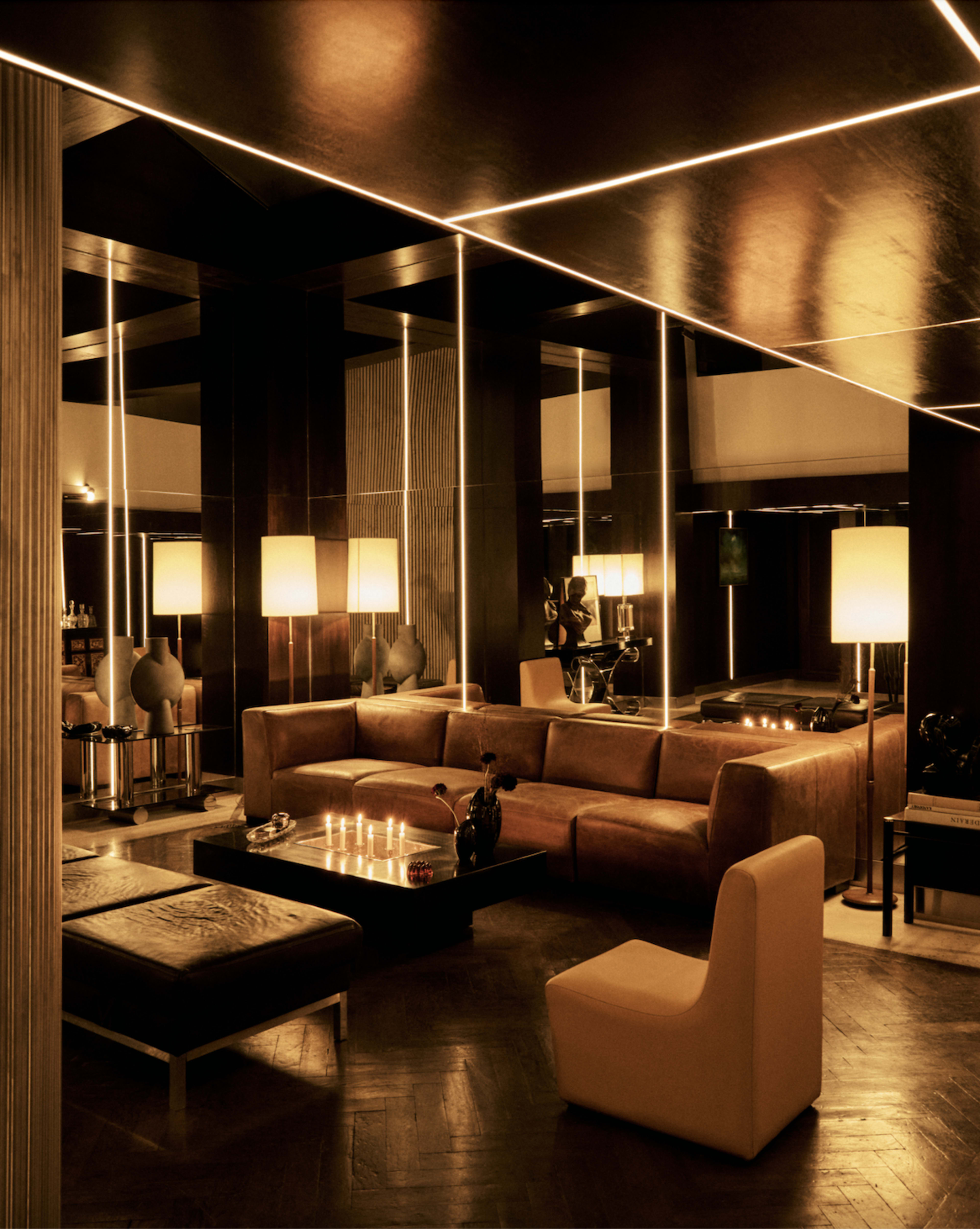 The image depicts a stylish, modern lounge with a brown leather sectional sofa, a black coffee table, and illuminated lamps, surrounded by mirrors and warm lighting.