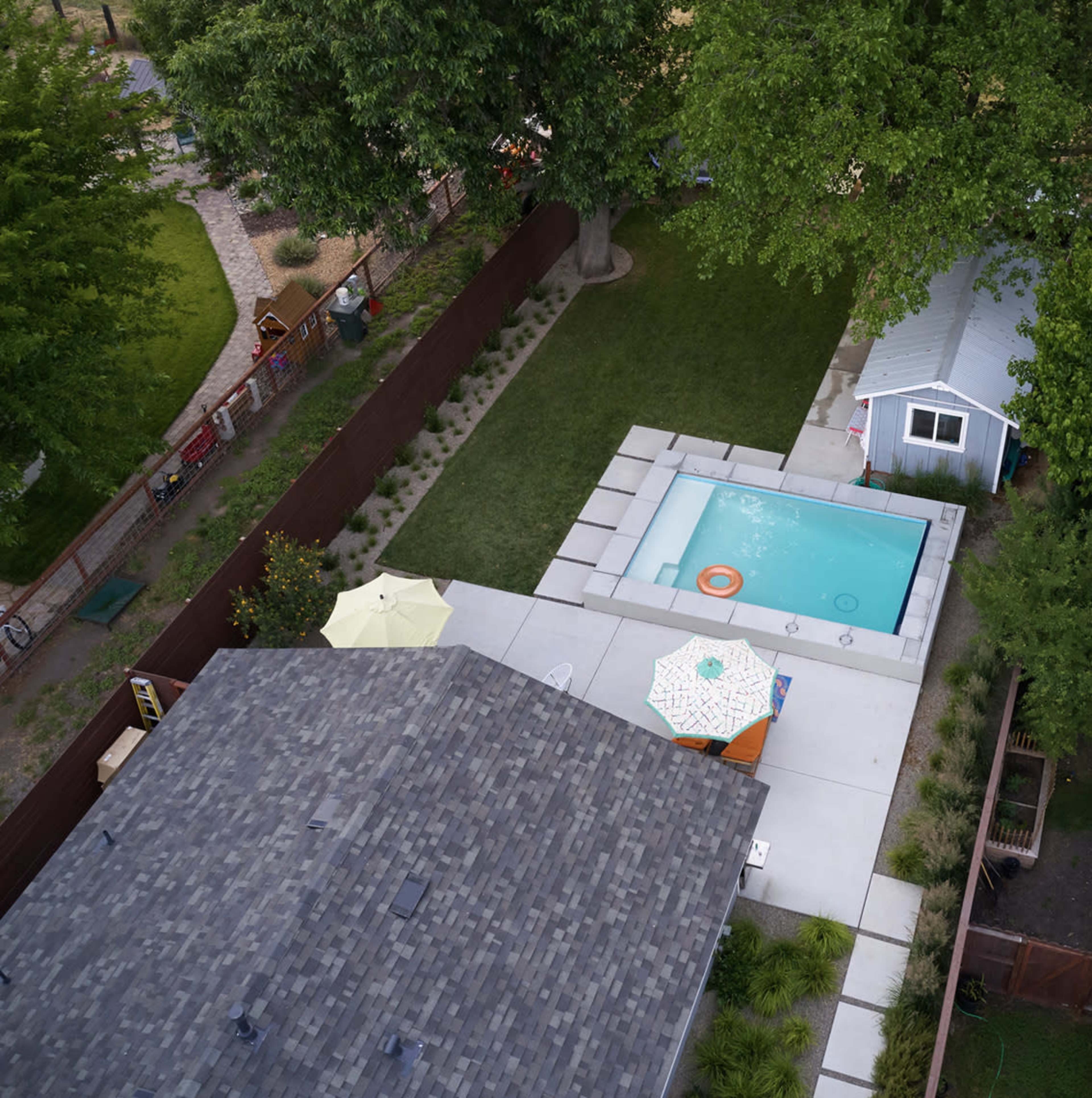 The image shows a backyard with a swimming pool surrounded by concrete, a small shed, and an area of grass bordered by a fence.