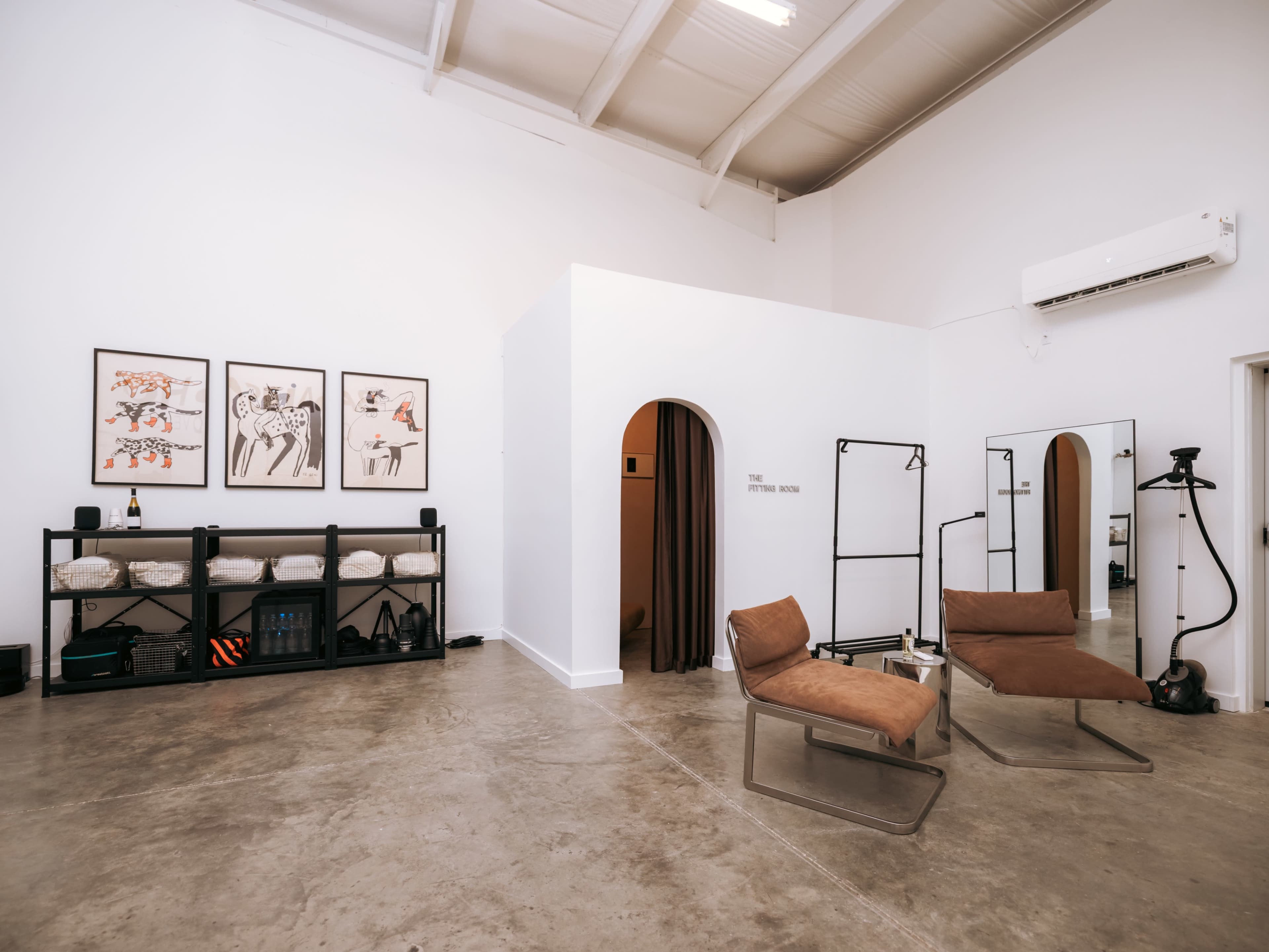 The image shows a modern interior space with two brown lounge chairs, a minimalist shelf with decorative items, framed artwork on the wall, and a partition leading to another room.