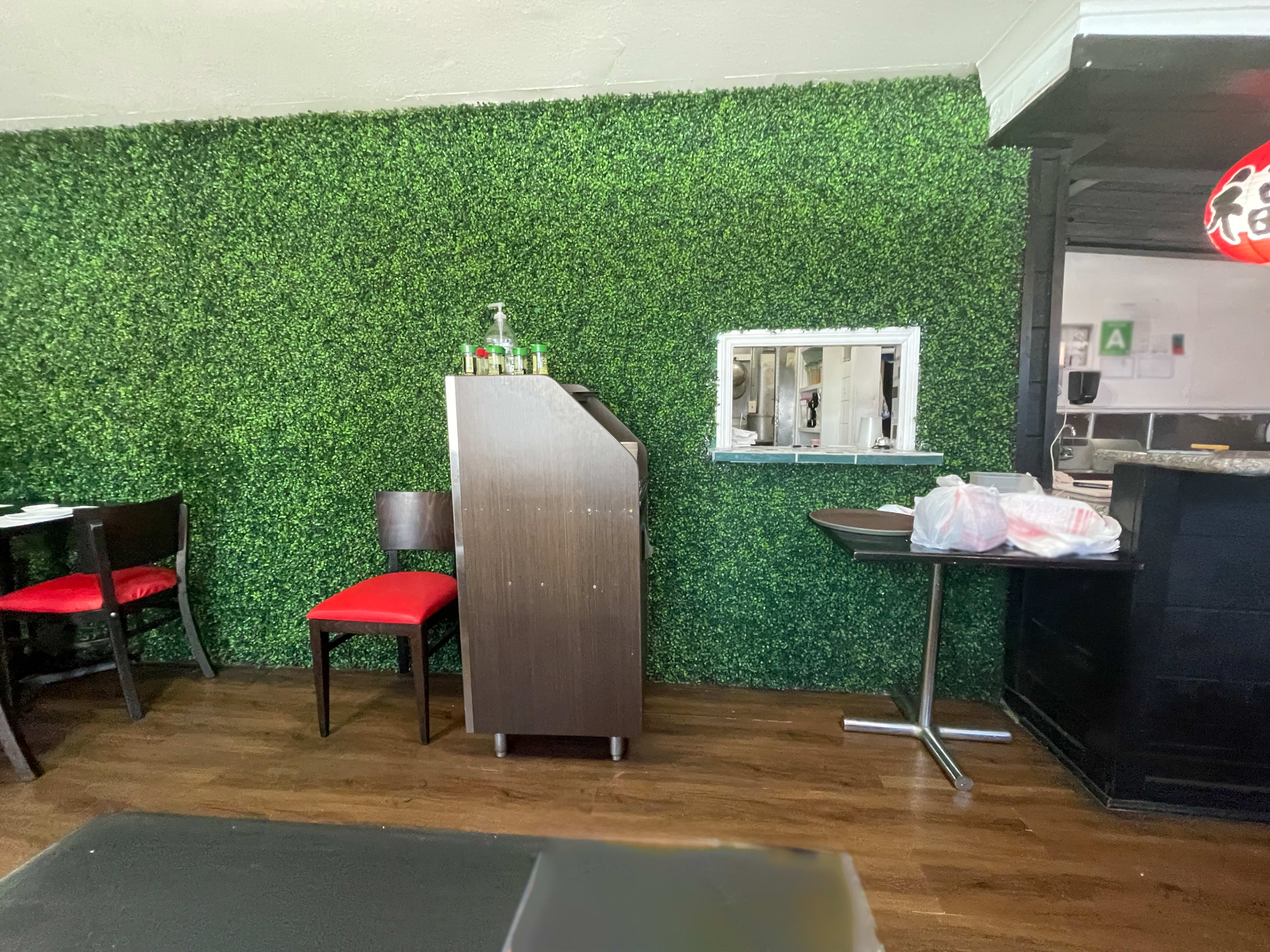 The image shows a restaurant interior featuring a green artificial hedge wall, a wooden podium, and a table set with a takeout bag.