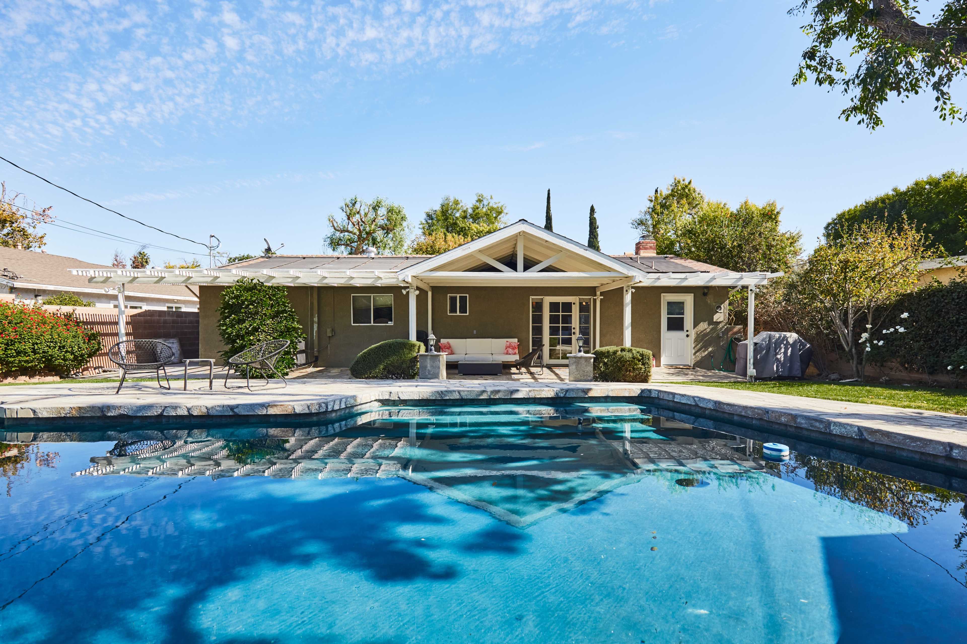 A backyard with a swimming pool in front of a single-story house, surrounded by trees and landscaped greenery.