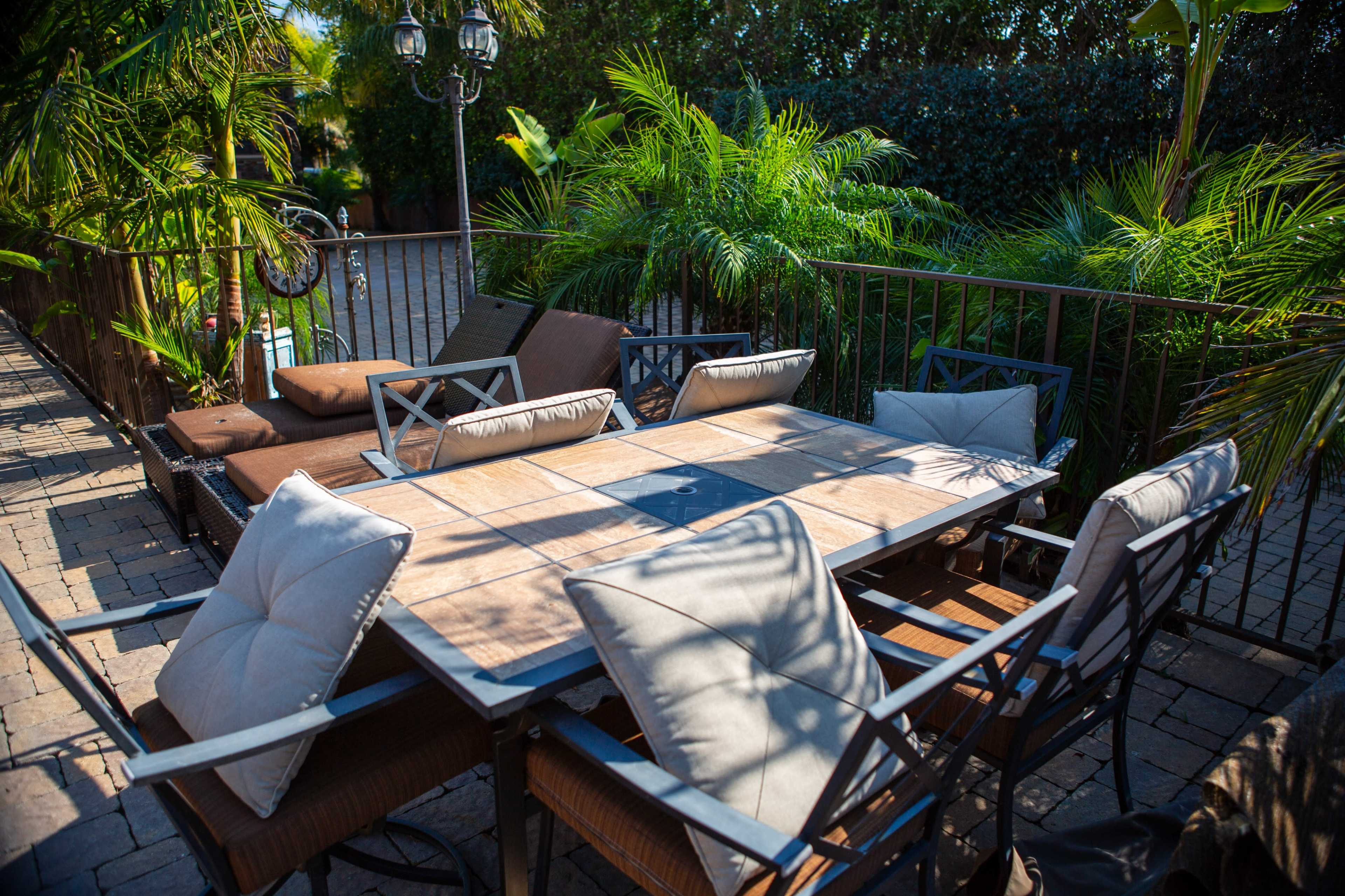 A rectangular outdoor dining table surrounded by cushioned metal chairs is situated on a stone patio, bordered by lush greenery.