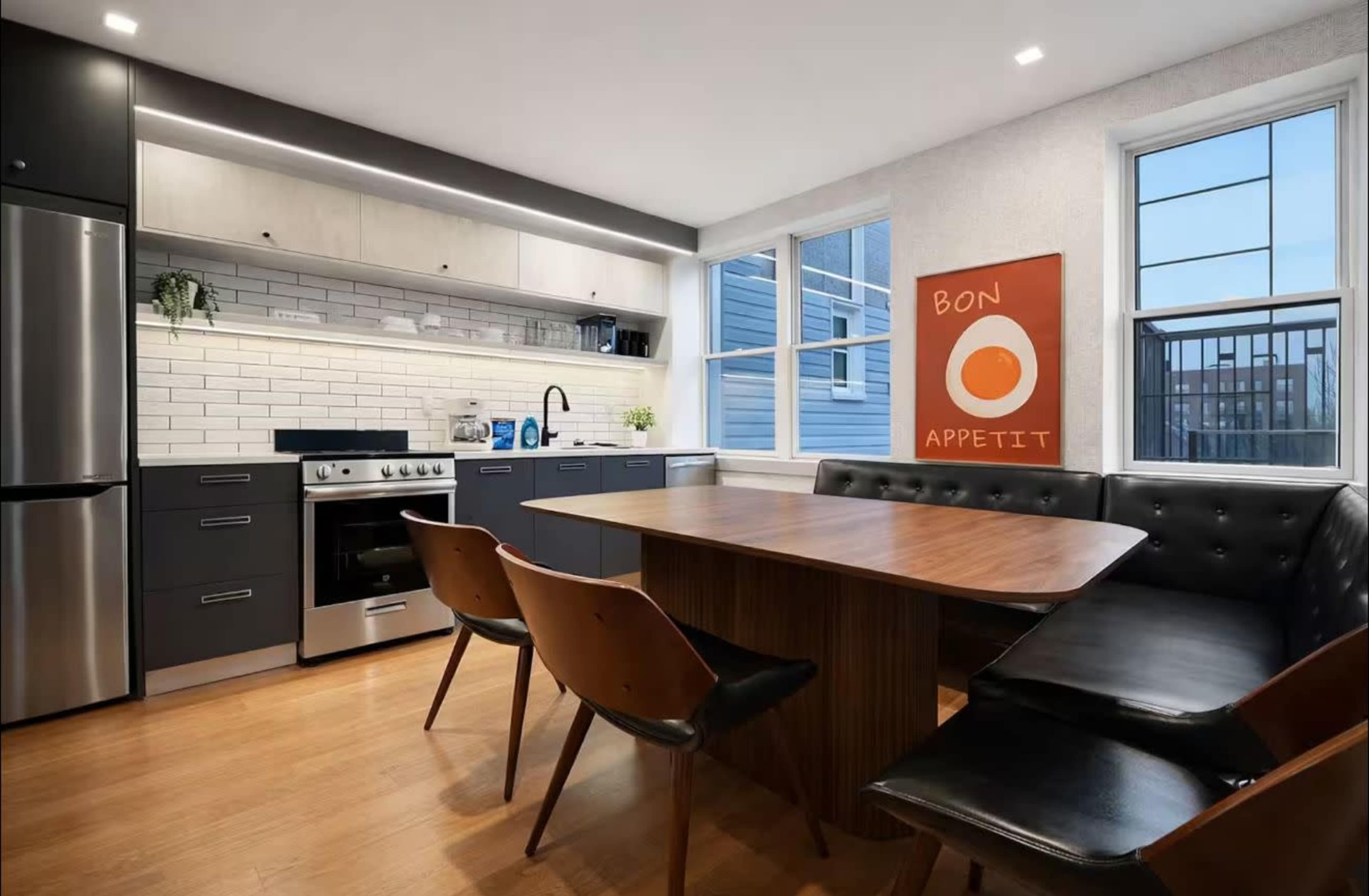 A modern kitchen features sleek cabinetry, a stainless steel oven, and a dining area with a wooden table and black leather seating.