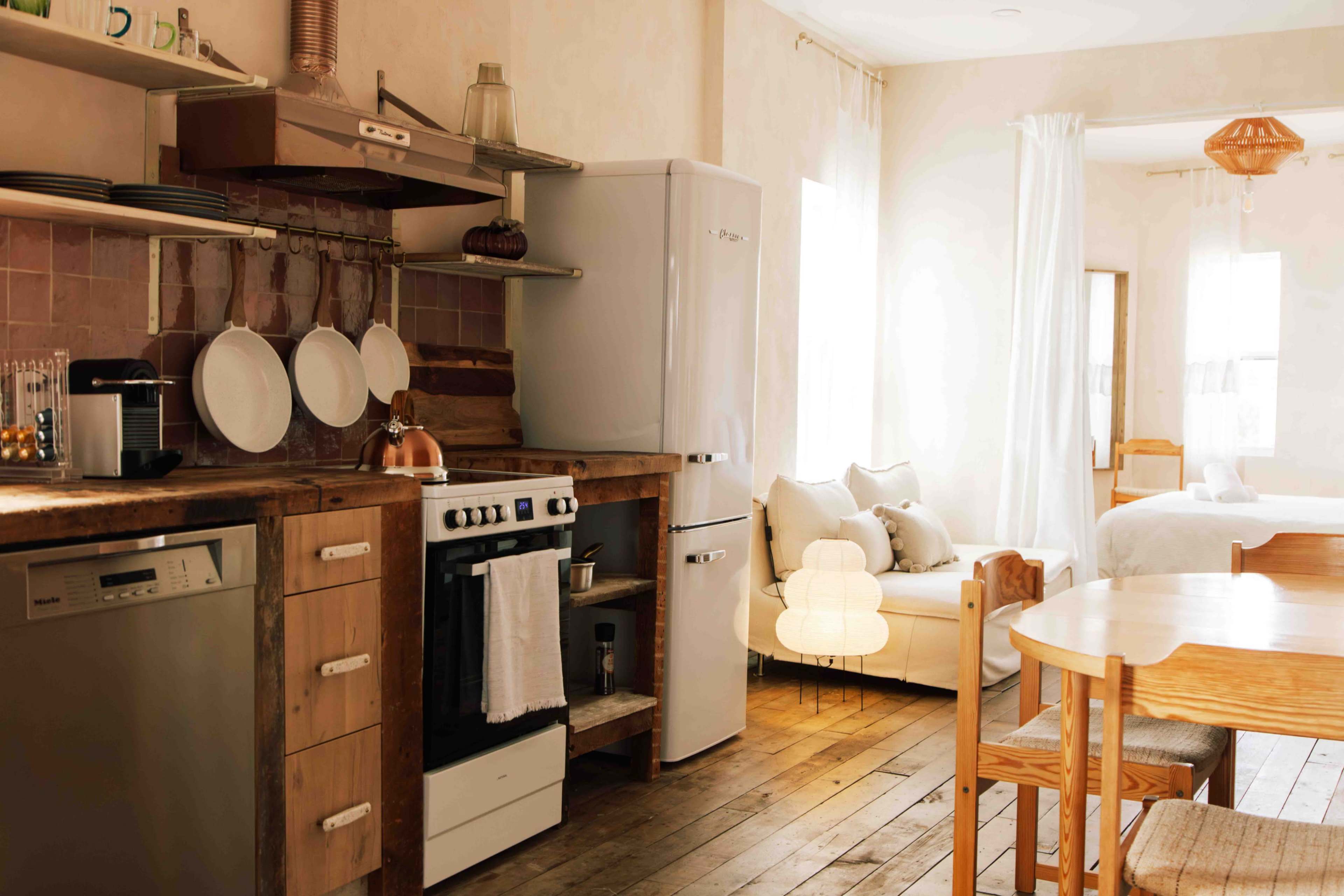 The image shows a modern kitchen area with a wooden counter, a white refrigerator, a stove, and a dining table, all set in a bright, airy space.