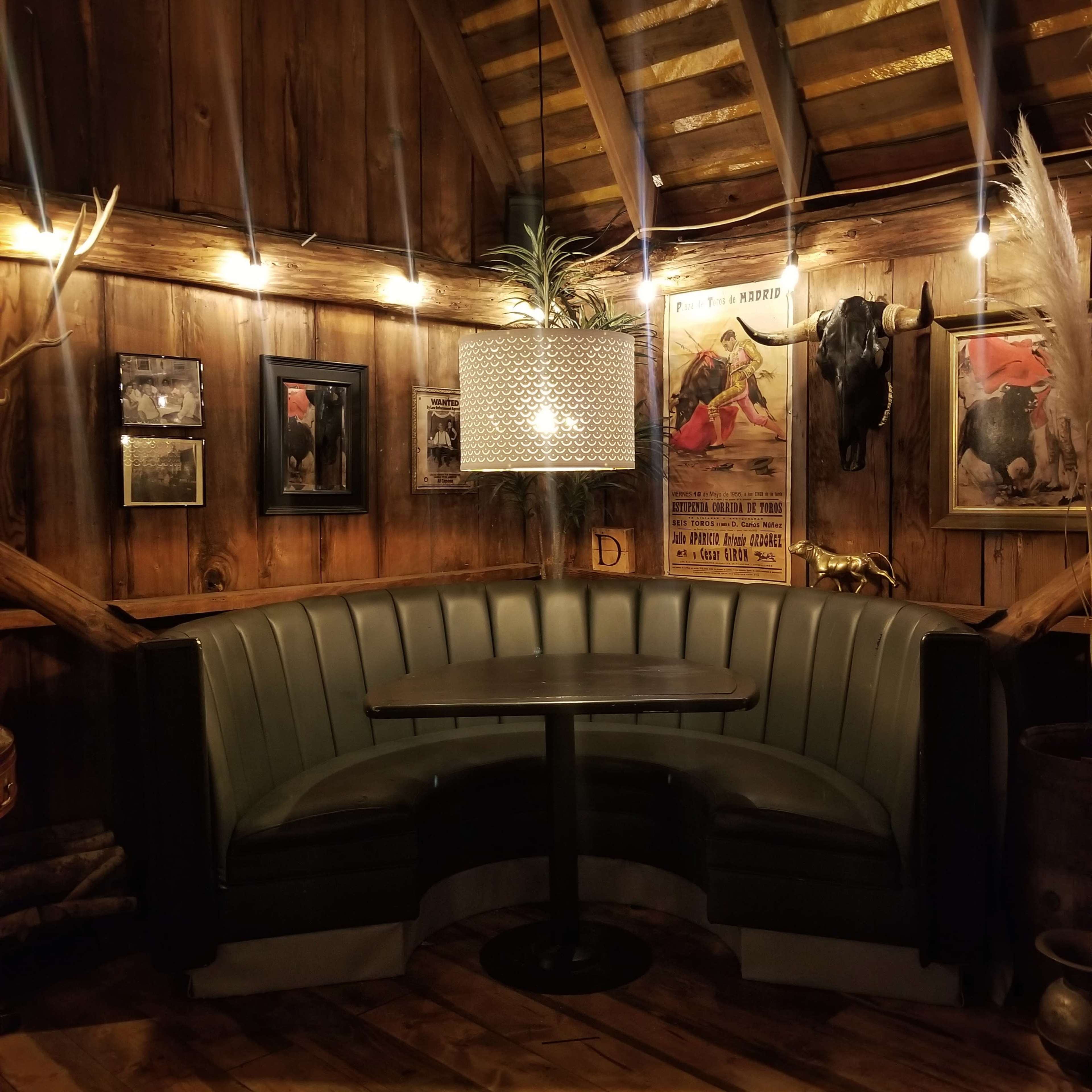 A curved leather booth with a round table is illuminated by a hanging light fixture in a rustic wooden room adorned with animal-themed decor and framed artwork.
