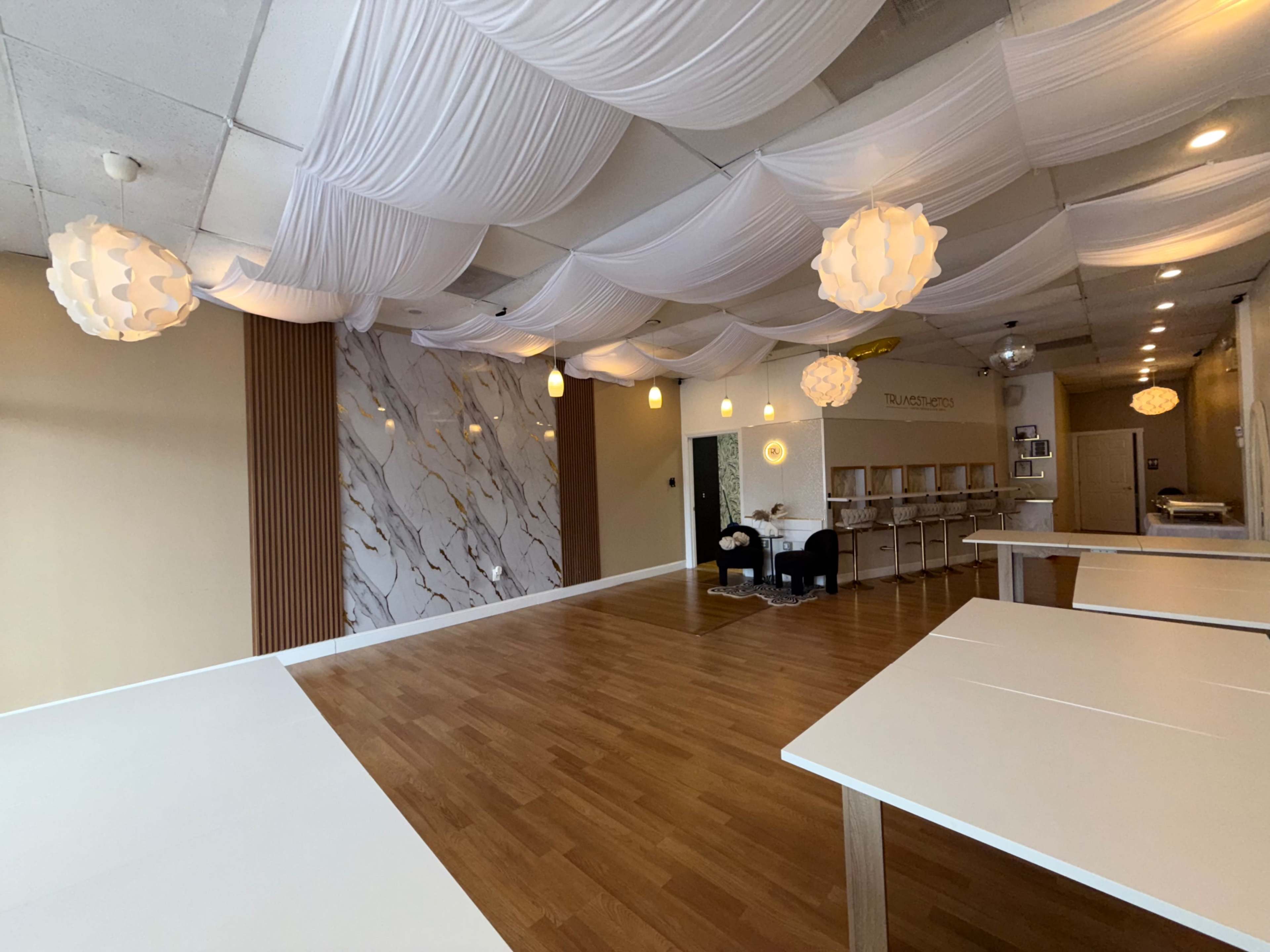 The image shows a spacious, well-lit room with white draped ceilings, wooden flooring, and a marble accent wall featuring modern light fixtures and furniture.