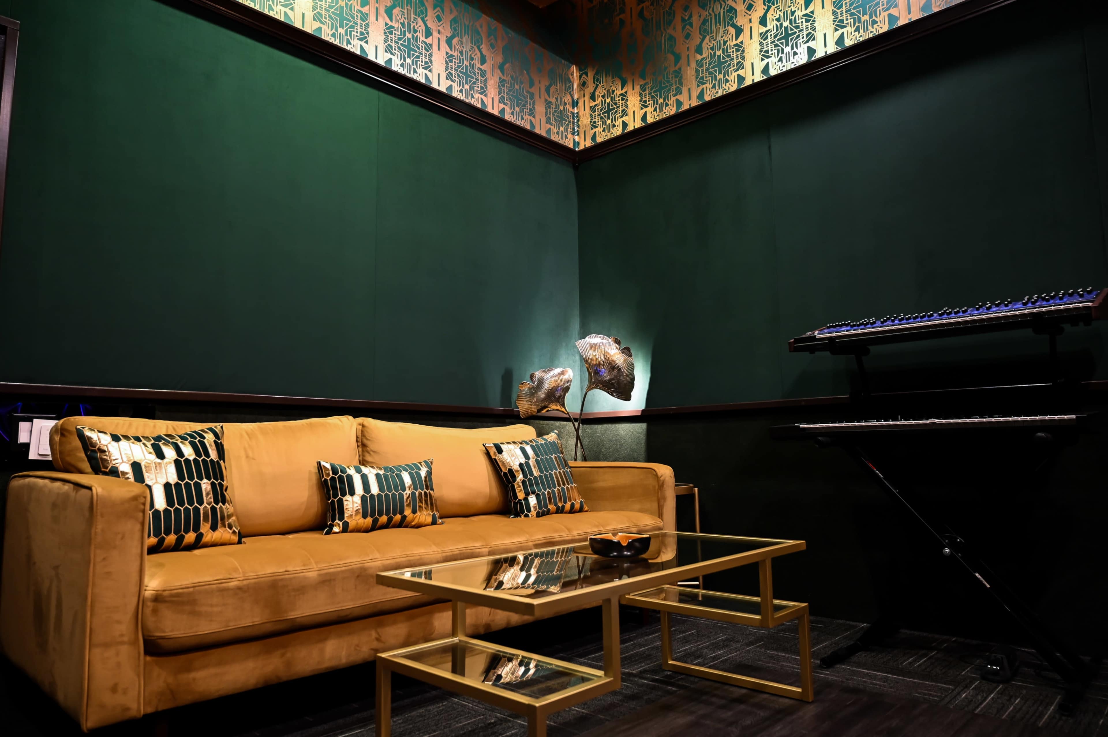 The image shows a cozy lounge area featuring a golden sofa with decorative pillows, a glass coffee table, and a keyboard on a stand against a dark green wall.
