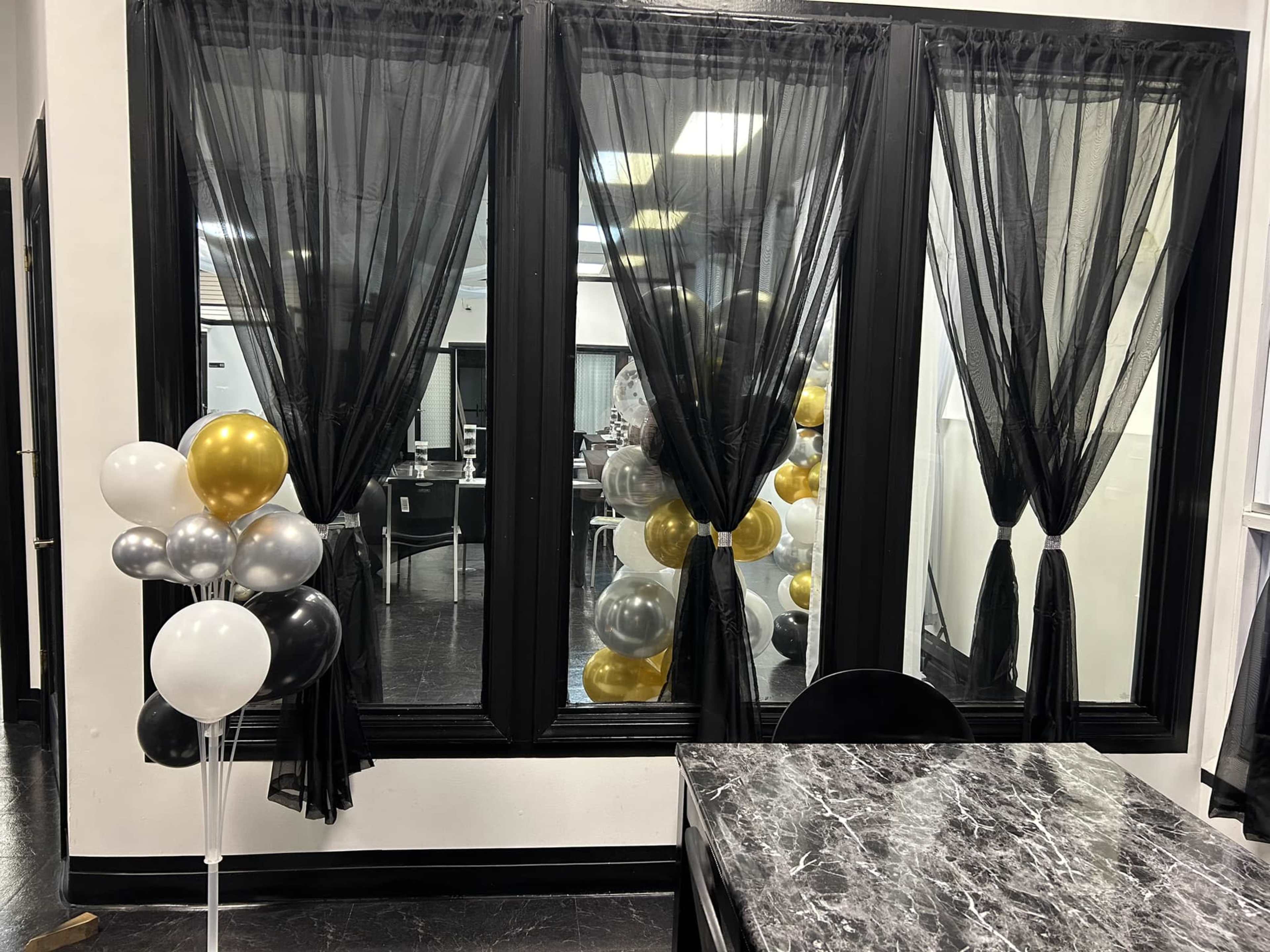 A room with large black-tinted windows draped in sheer black curtains, alongside a cluster of balloons in black, gold, silver, and white near a marble-topped table.