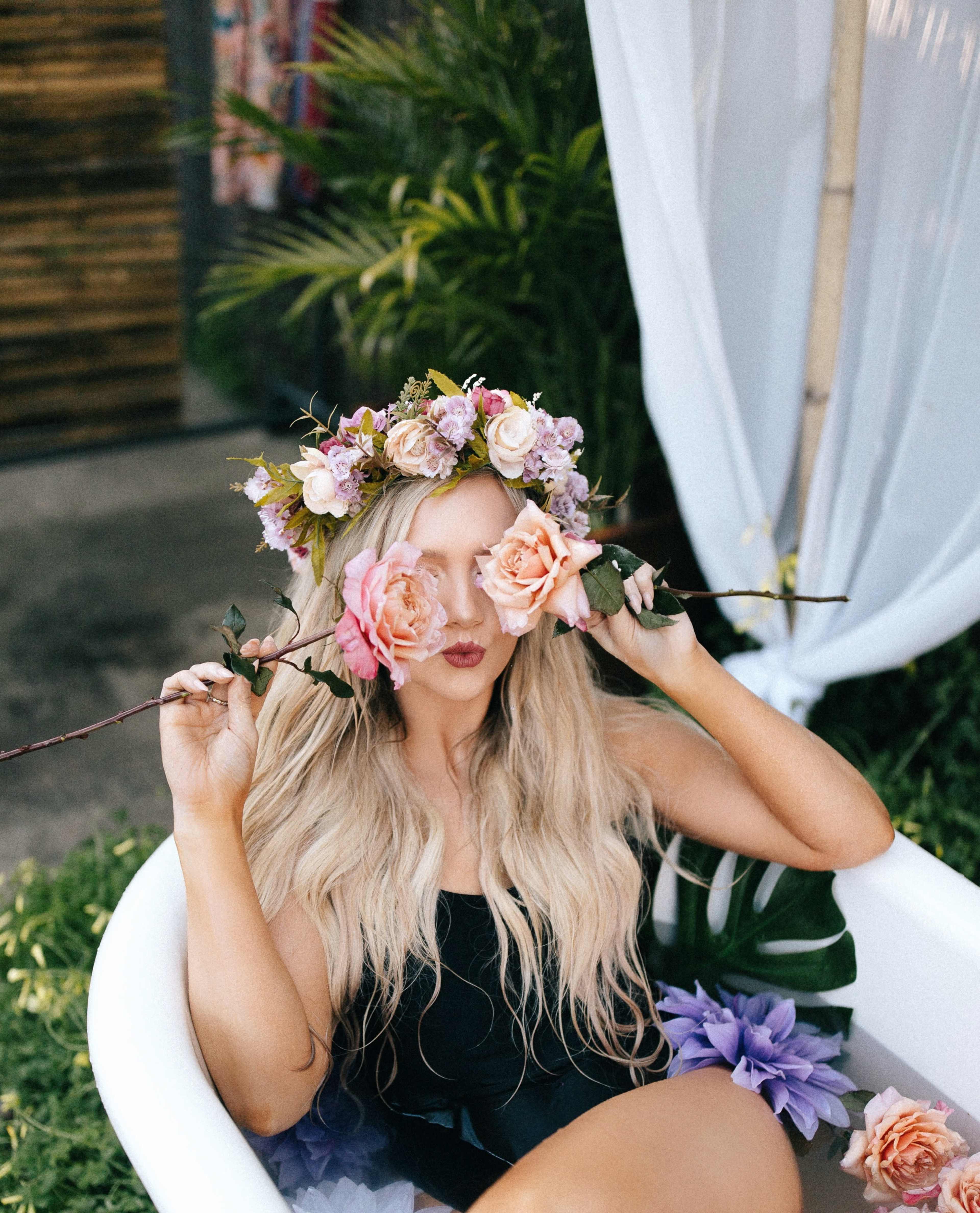 A person wearing a floral crown sits in a bathtub surrounded by greenery and flowers.