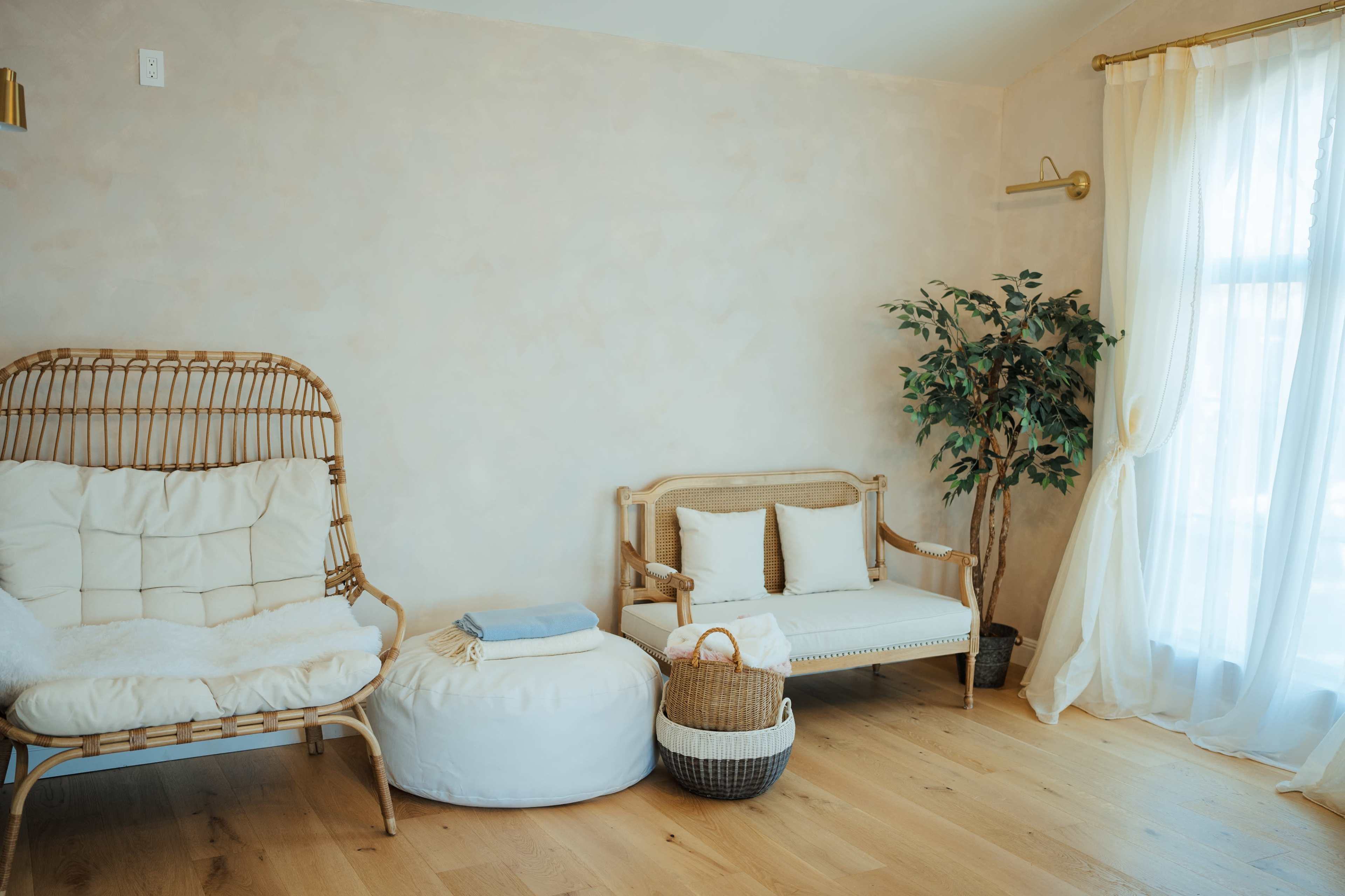 A cozy living space with a rattan chair, a small sofa, a plant, and a pouf, all arranged on wooden flooring by a window with light curtains.